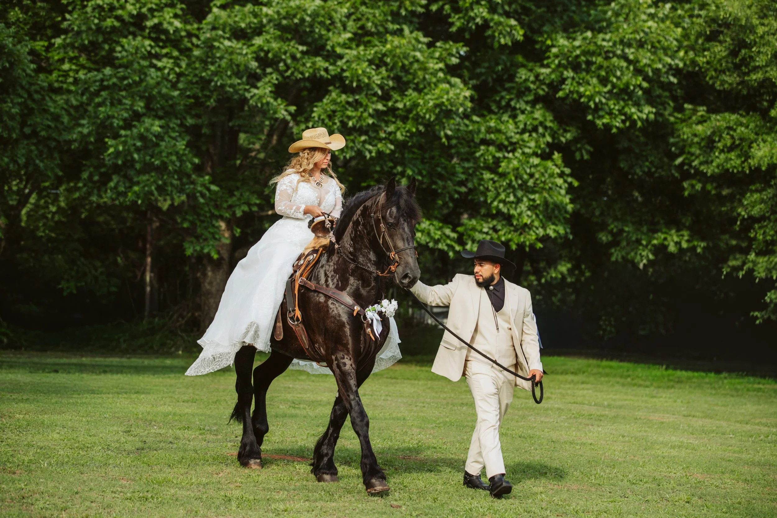 A woman in a white lace wedding dress and wide-brimmed straw hat riding a black horse, accompanied by a man in a beige suit, black hat, and holding the horse's reins, in a green outdoor setting.