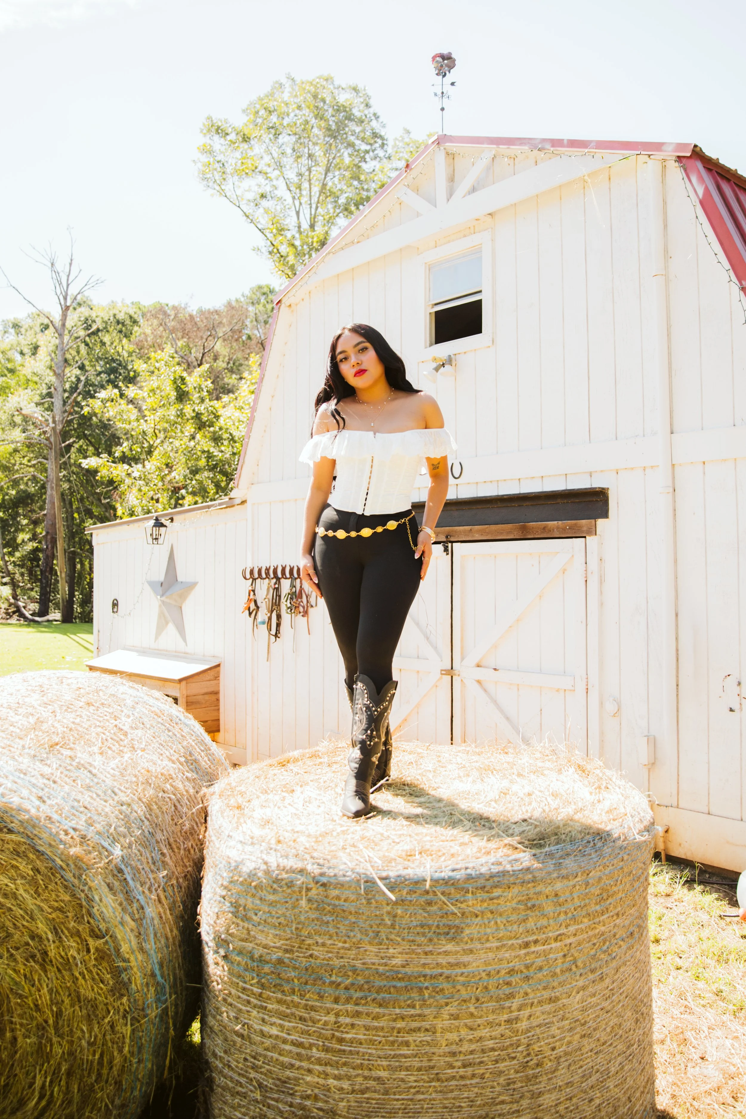 A woman with long dark hair in a white off-the-shoulder top and black pants standing on hay bales in front of a white barn with string lights on a sunny day.