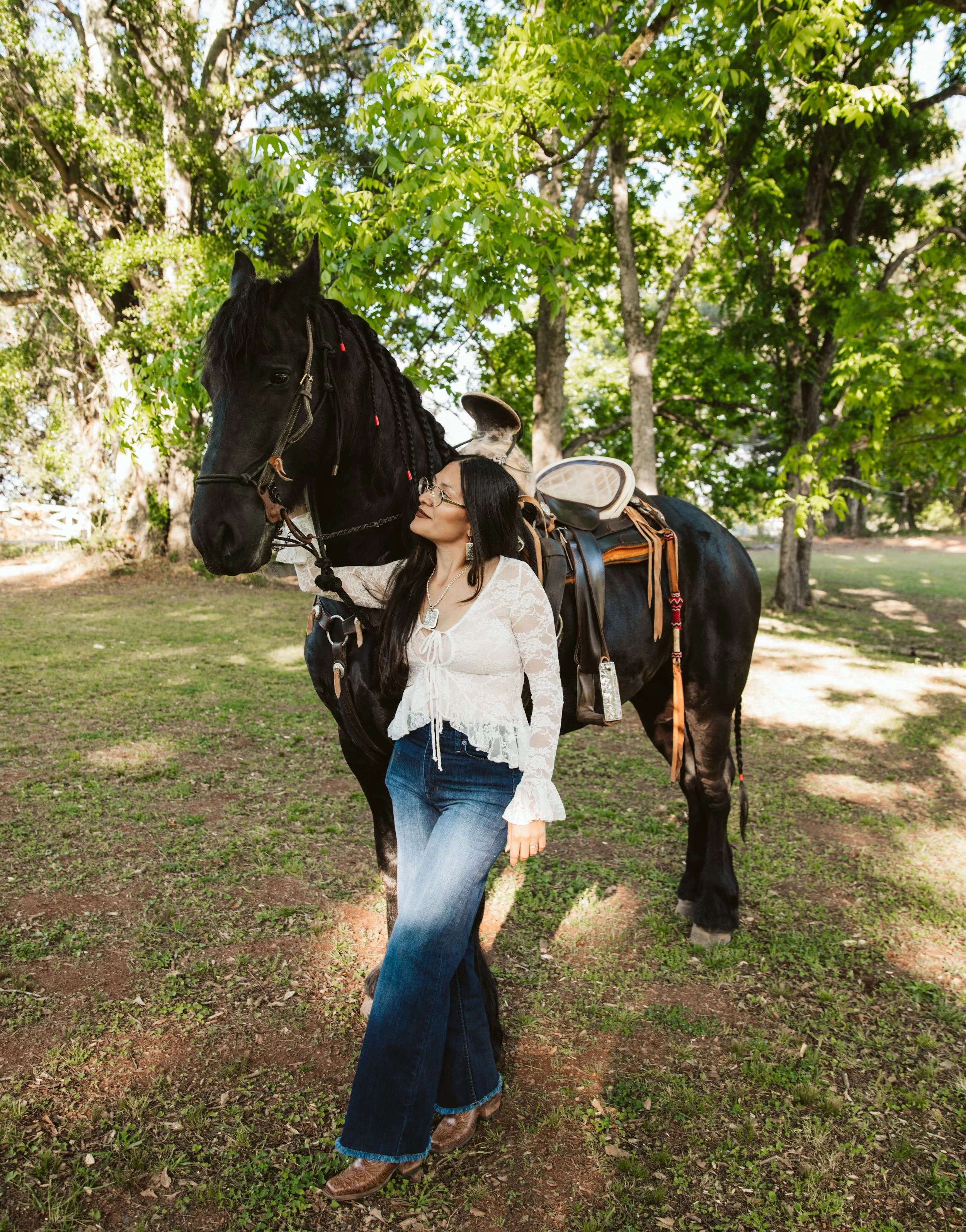 A woman with long dark hair, glasses, and cowboy boots is standing next to a black horse in a wooded area. The woman is wearing a white lace top and blue jeans, and the horse is saddled with gear, in a sunlit forest with green leaves.