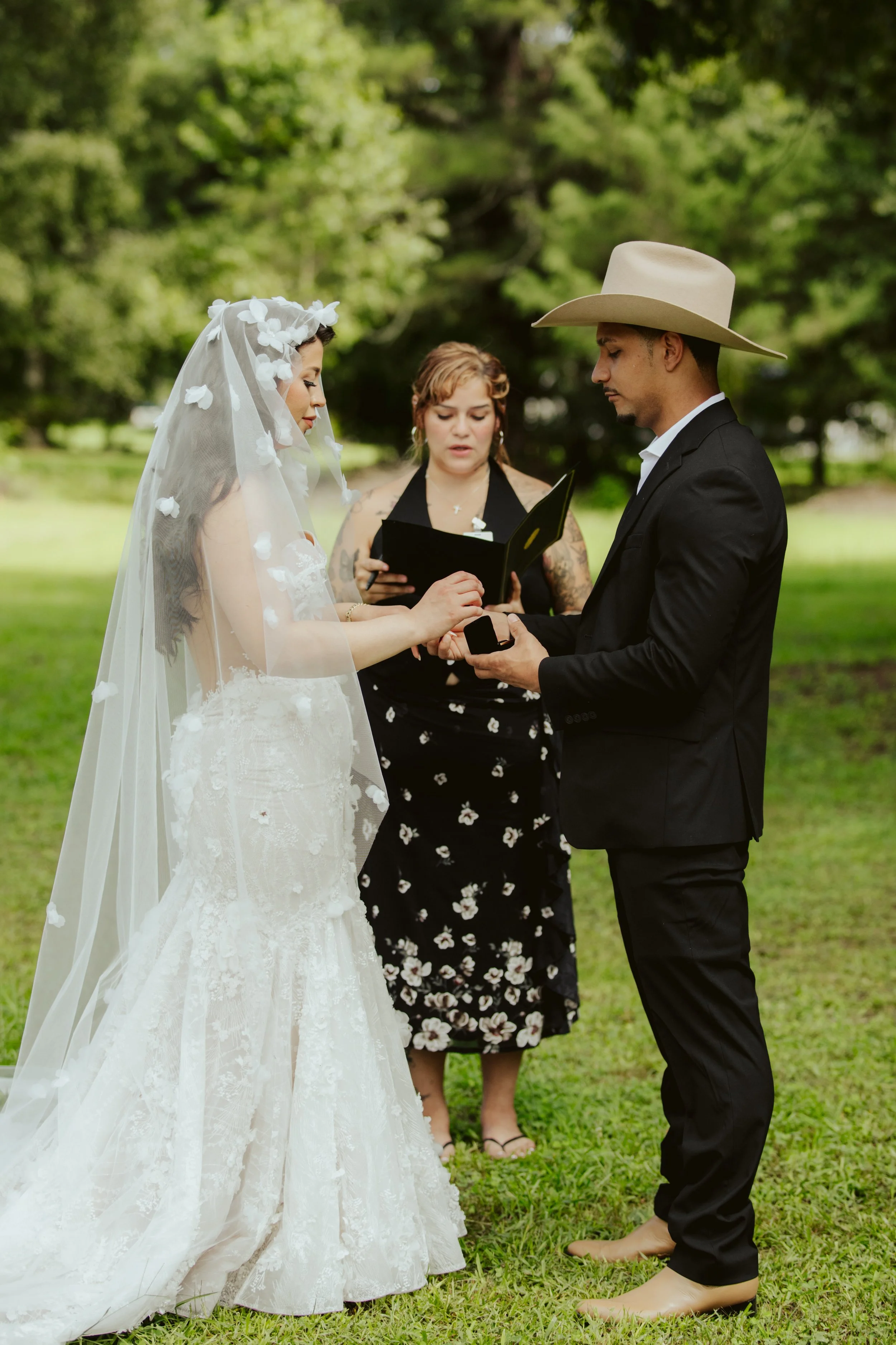 A couple getting married outdoors, with a woman officiant in the background, in front of green trees. The bride wears a white wedding gown and veil with floral details, and the groom wears a black suit and cowboy hat, holding a ring box.