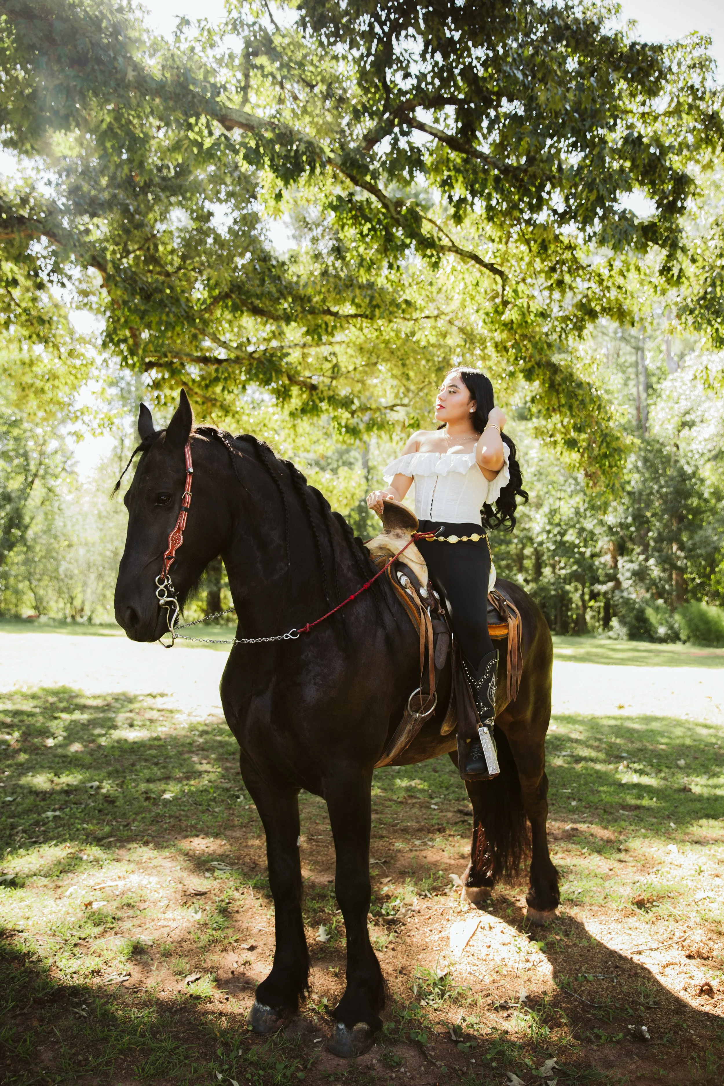 A woman with long dark hair riding a black horse in a park with green trees, wearing a white off-the-shoulder top and black pants.