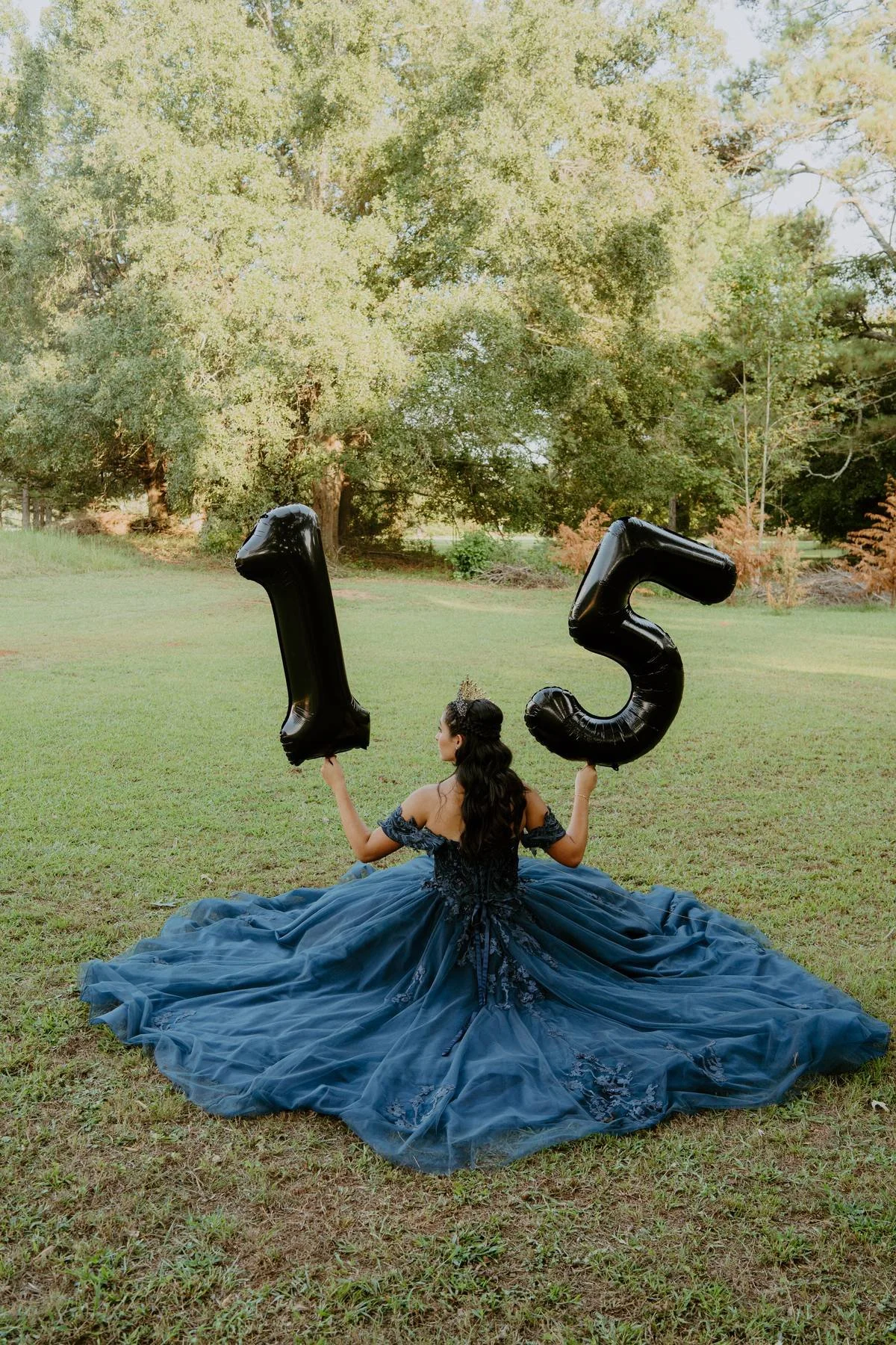 A woman in a dark blue, formal gown with intricate lace details is sitting on a grassy field, holding two black balloons shaped as the numbers 1 and 5, indicating a 15th birthday celebration. She is sitting outdoors with a background of trees and greenery.
