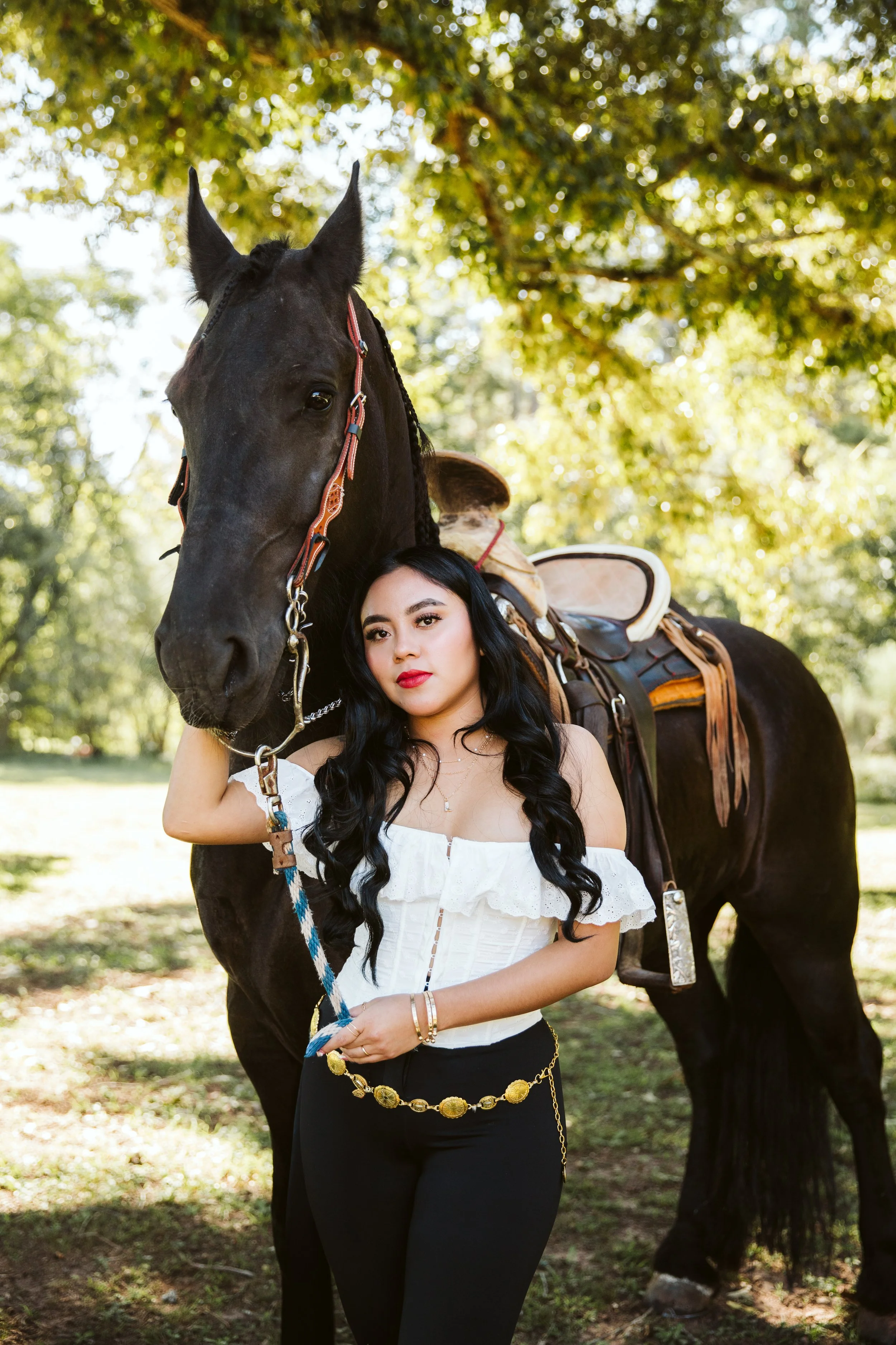 A woman with long dark hair and red lipstick standing outdoors in sunlight, holding a blue and white lead attached to a black horse that has a brown saddle and bridle. The background shows green trees and a sunny day.
