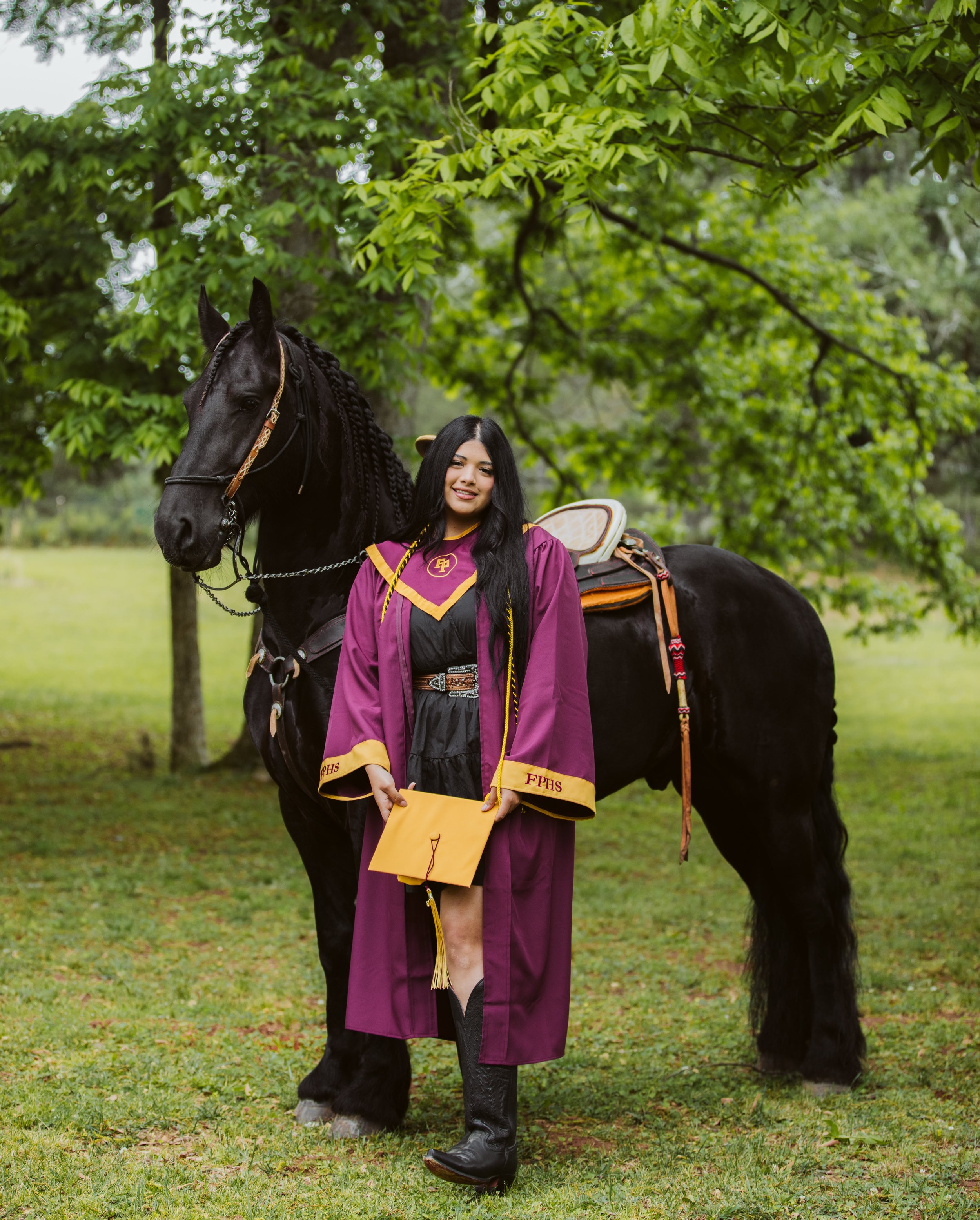 A young woman in graduation gown holding diploma, standing next to a black horse on a grassy area with trees in the background.