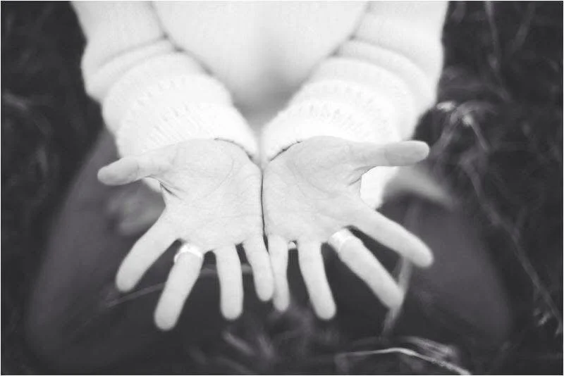 A pair of outstretched hands with palms facing up, in front of a person wearing a white sweater. The photo is black and white.
