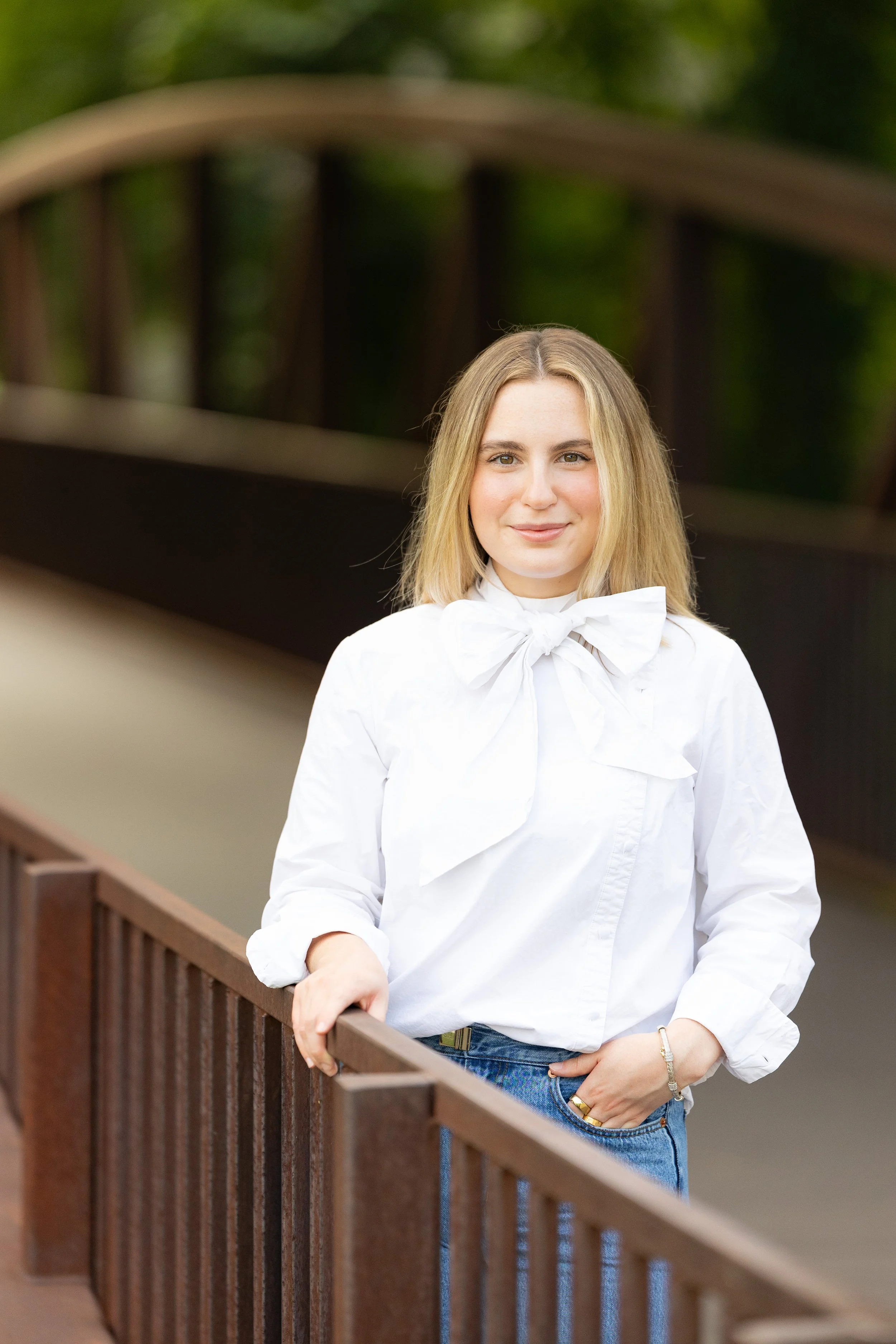 A young woman with blonde hair, wearing a white blouse with a bow and blue jeans, standing on a wooden bridge with green trees in the background.