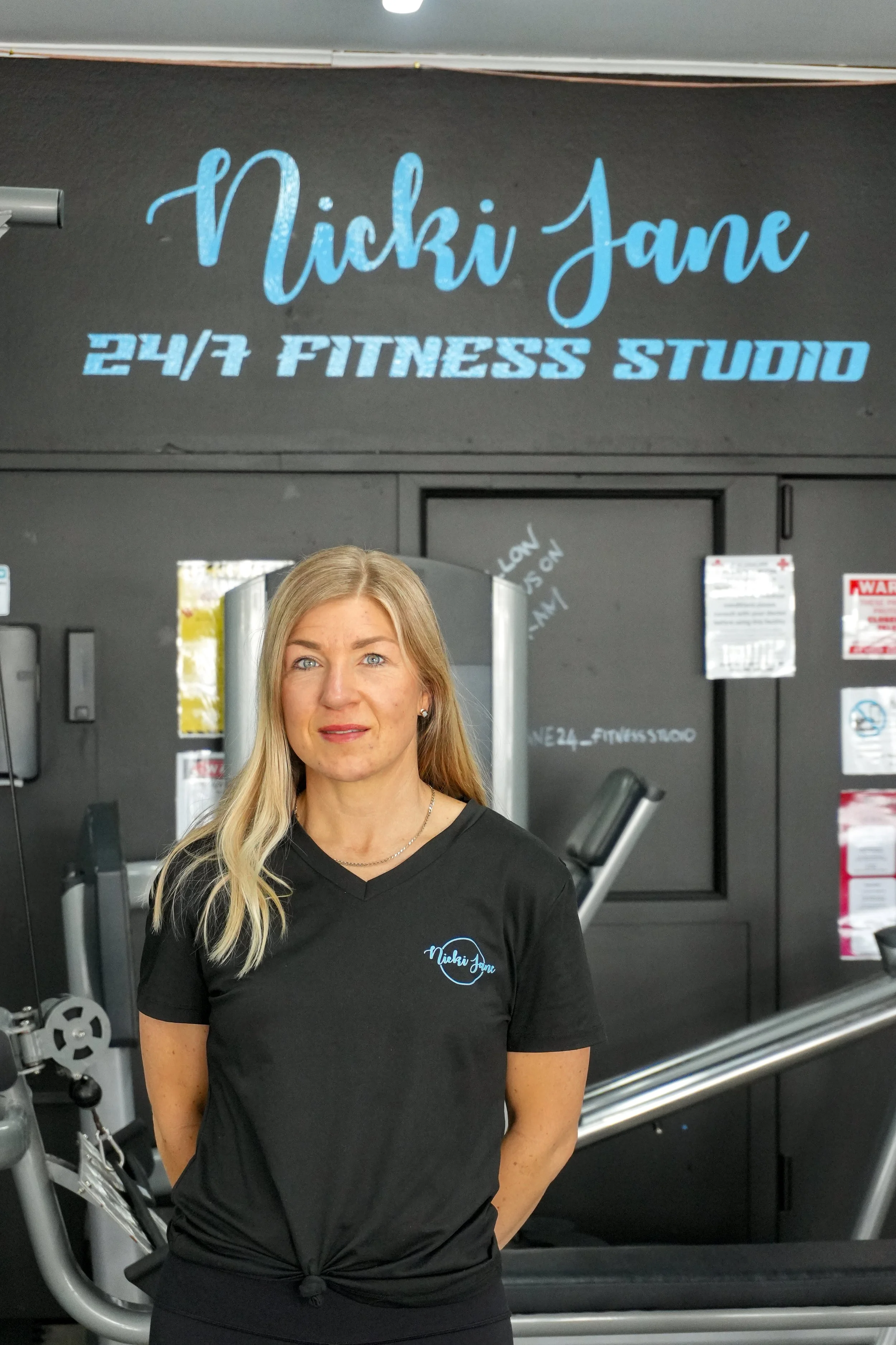 A woman with long blonde hair wearing a black T-shirt with a logo that says 'Nicki Jane' stands in a gym in front of fitness equipment. The background features a black wall with a sign that says 'Nicki Jane 24/7 Fitness Studio' in blue.