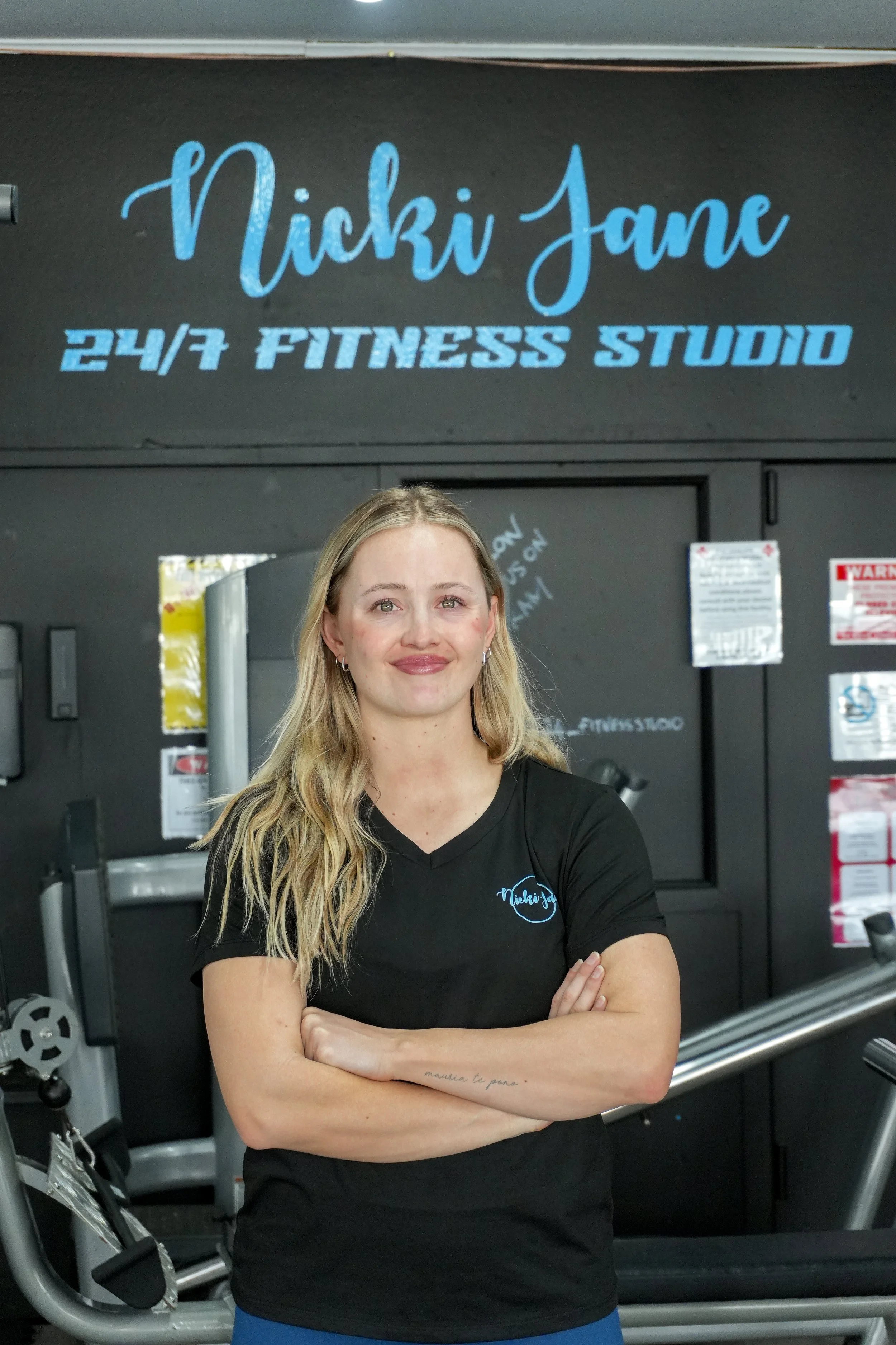 A woman with long blonde hair and tattoos on her arm, smiling with arms crossed, standing in a gym with a sign above that reads 'Nicki Jane 24/7 Fitness Studio'.
