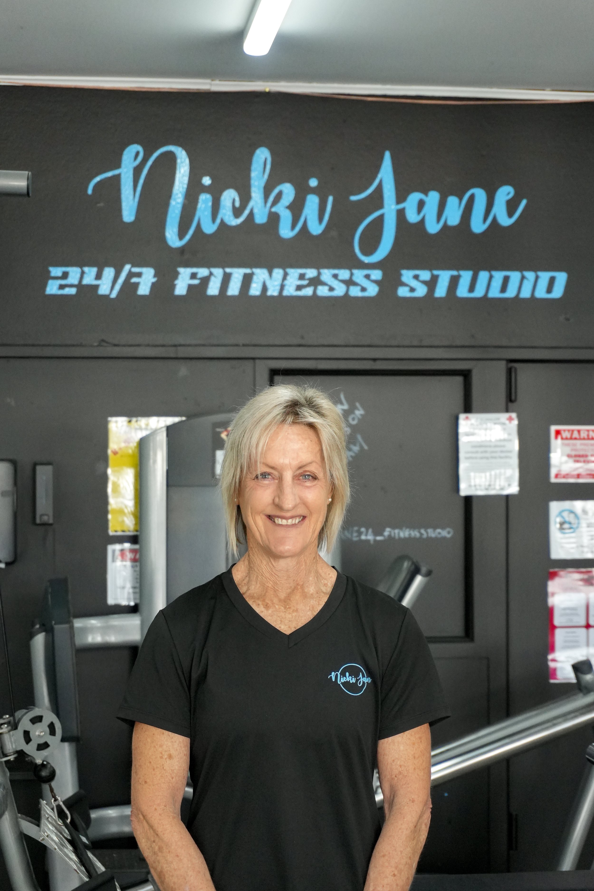 Smiling woman with short blonde hair wearing a black T-shirt with the name 'Nicky Jane' in blue on the front, standing in a gym with exercise equipment and a sign that reads '24/7 Fitness Studio' in the background.