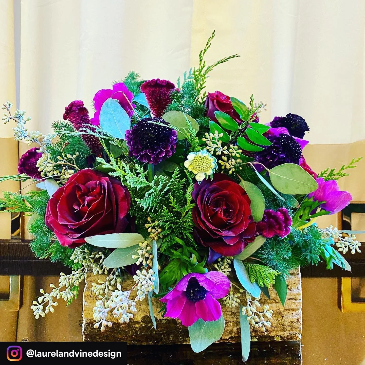 Colorful floral arrangement with red roses, purple and pink flowers, green foliage, and white accents on a table with a beige background.