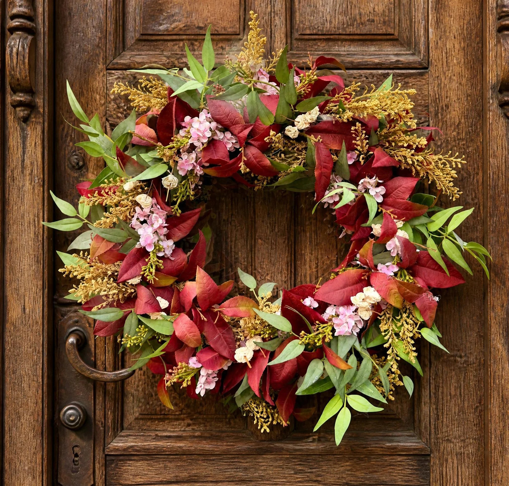 A floral arrangement of pink and white flowers, red and green leaves, and yellow accents hanging on a wooden door with a vintage handle.