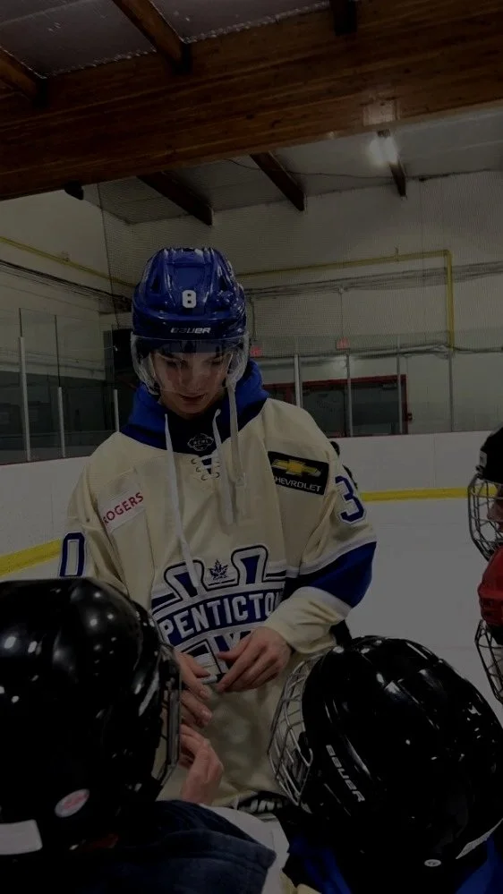 Hockey player in beige and blue jersey with the number 3, wearing a blue helmet, on an ice rink, surrounded by young players with black helmets, in a practice or team meeting.