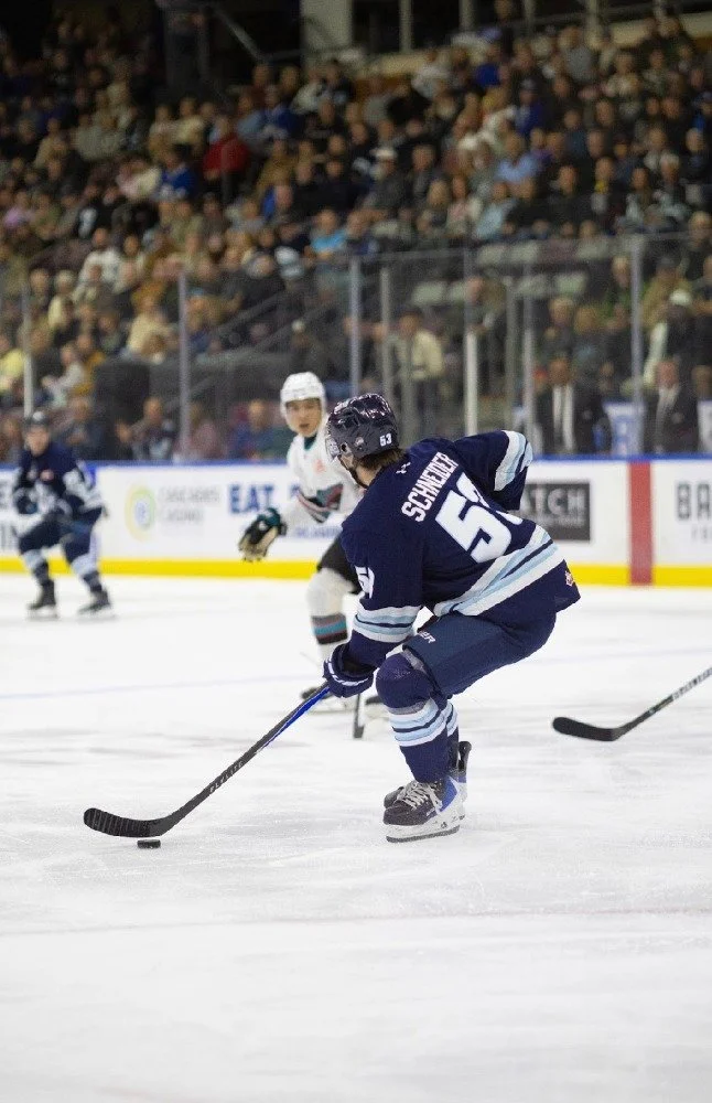 Ice hockey player in a navy blue uniform handling the puck during a game at an indoor arena with spectators in the background.