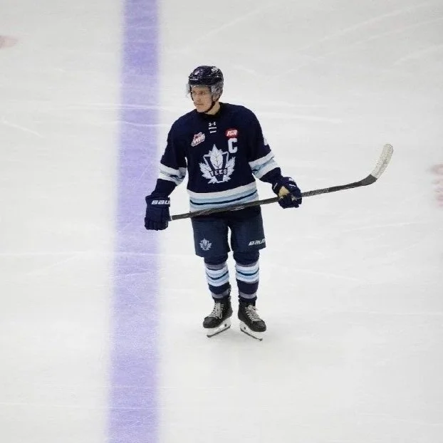Hockey player standing on ice hockey rink with stick, wearing a dark jersey with a maple leaf logo and a captain's 'C', black helmet, gloves, and skates.