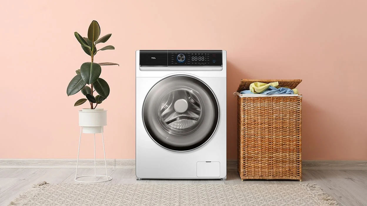 A laundry room with a washing machine, a potted plant, and a basket with clothes against a peach wall.