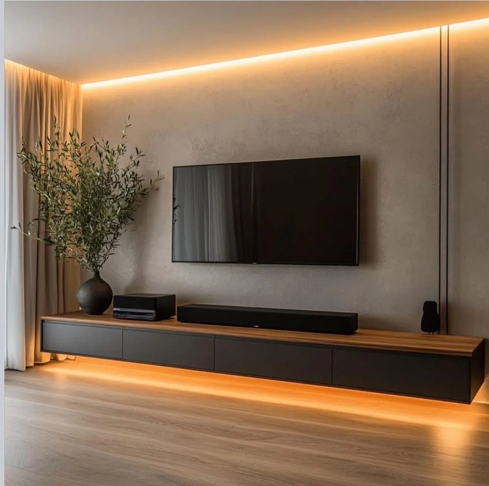 Modern living room with a large flat-screen TV mounted on the wall, a sleek black entertainment unit below, a tall potted plant on the left, and ambient lighting along the ceiling and under the unit.