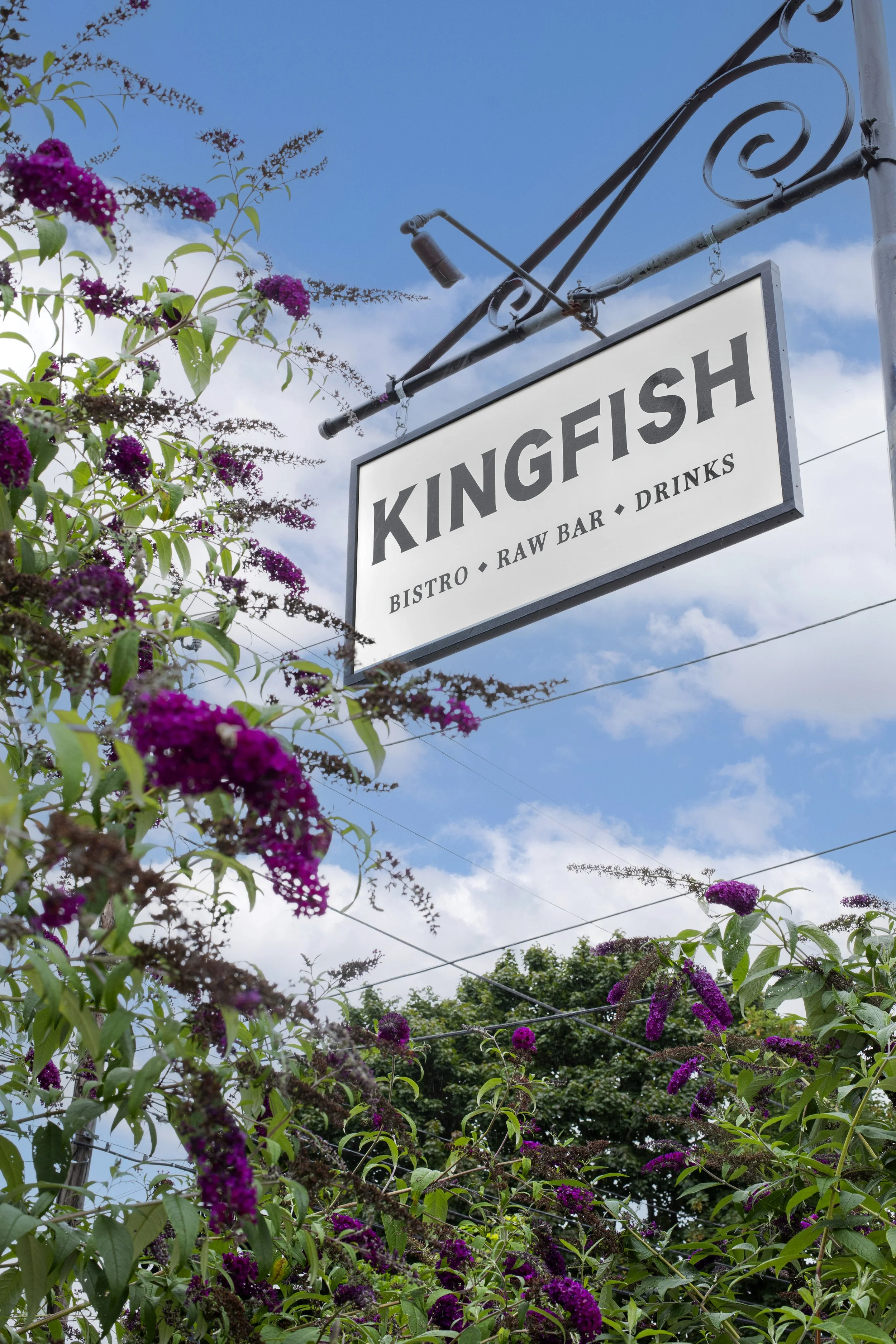 Sign for Kingfish Bistro, a restaurant and bar, with purple flowering plants and trees under a partly cloudy sky.