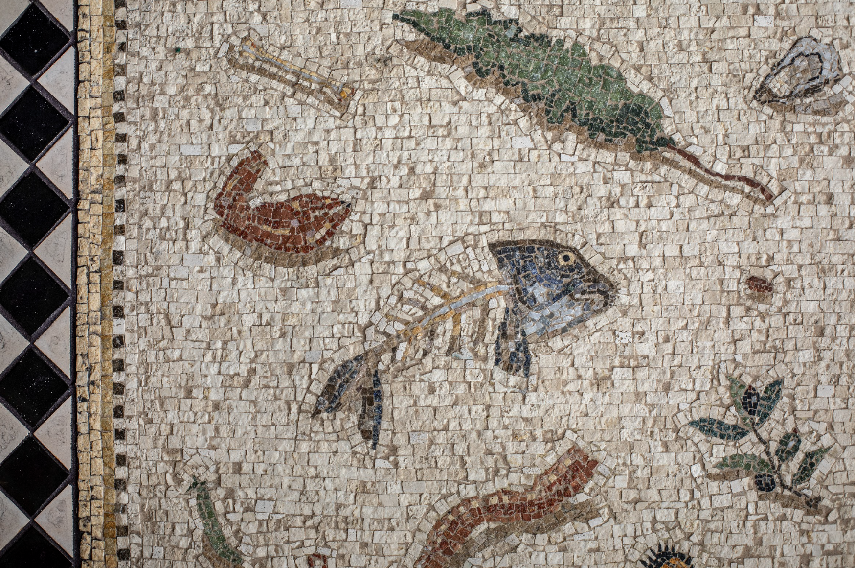 Close-up of a mosaic depicting fish, leaves, and abstract shapes made with small tiles on a wall.