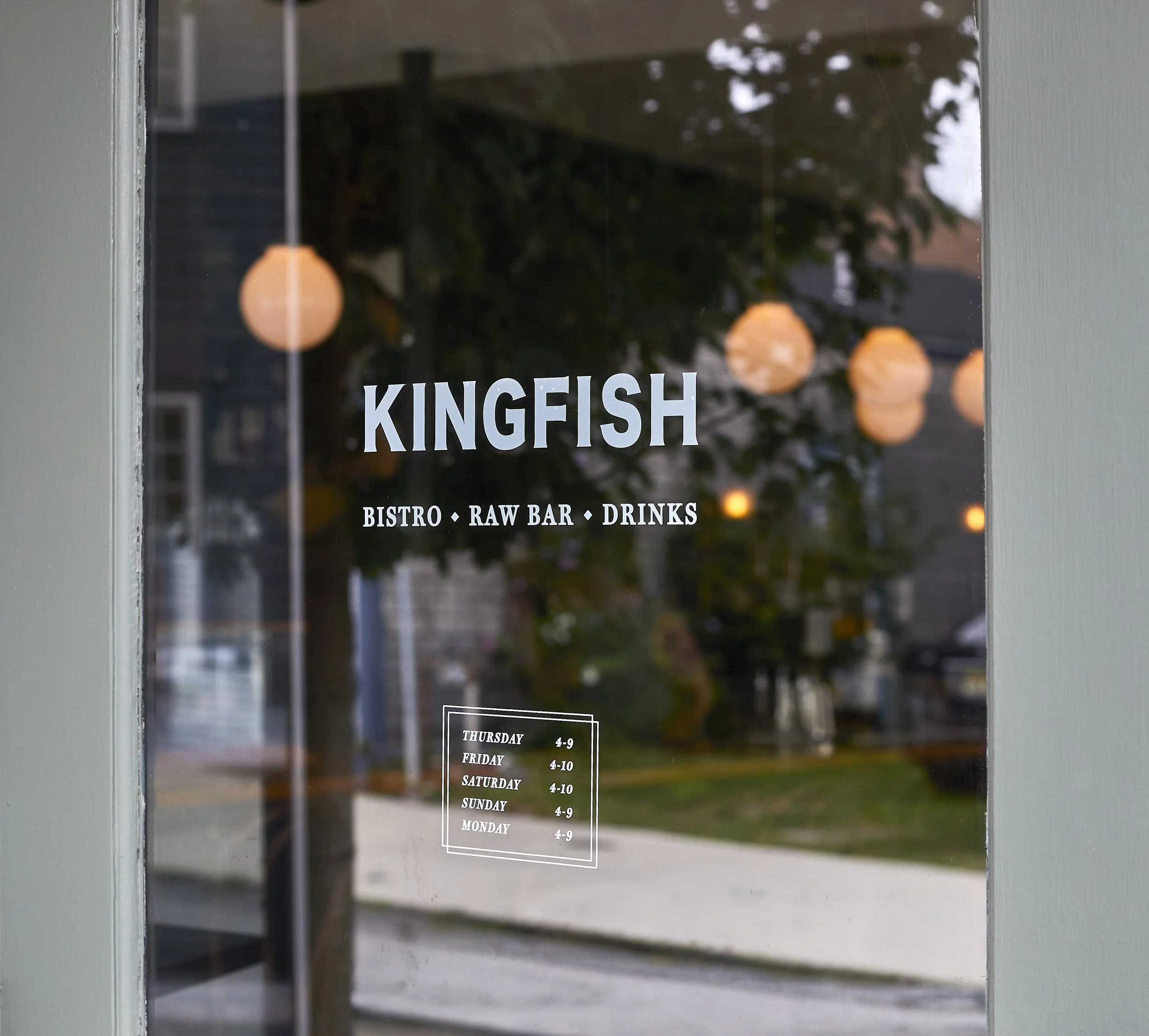 View through a glass door showing the name 'KINGFISH' and operating hours for each day of the week written in white text. Reflections of hanging lights and a tree outside are visible in the glass.