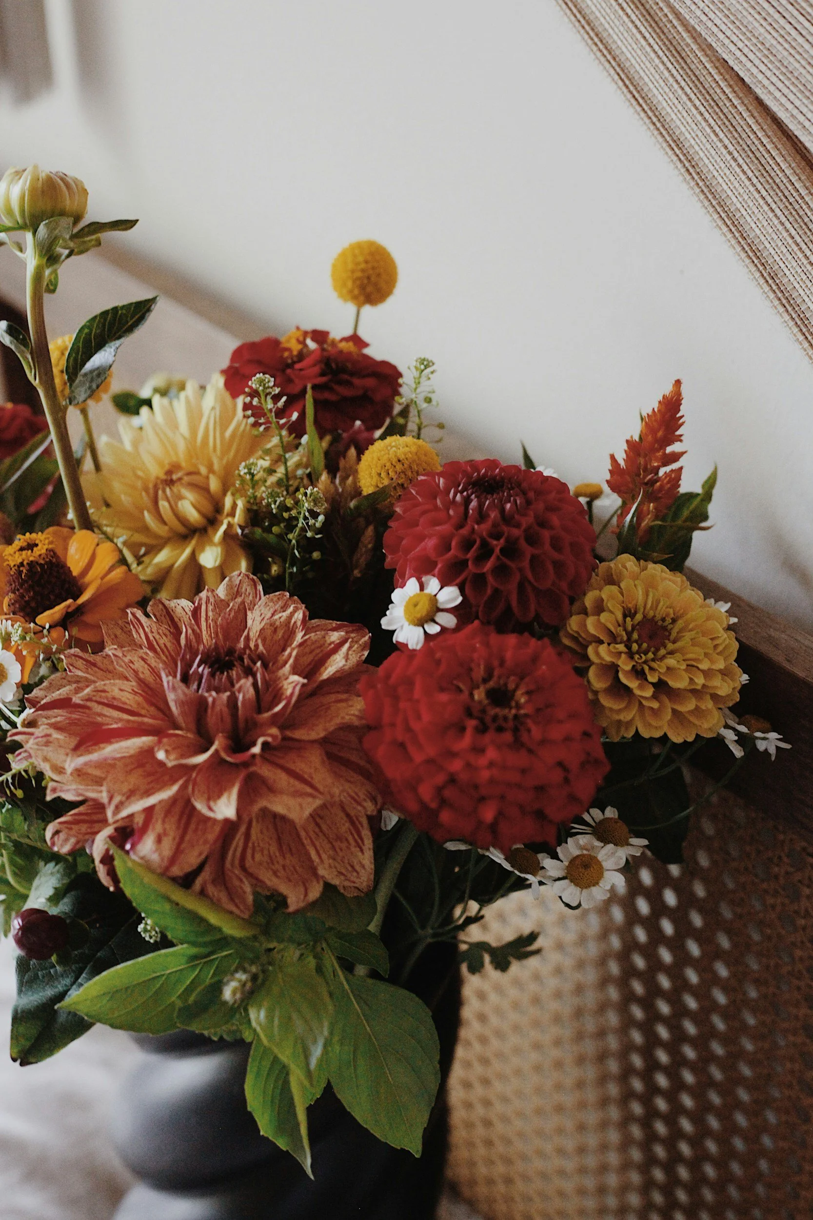 A colorful bouquet of various flowers including daisies, dahlias, and other blooms with green leaves in a black vase, placed on a surface next to a woven mat.