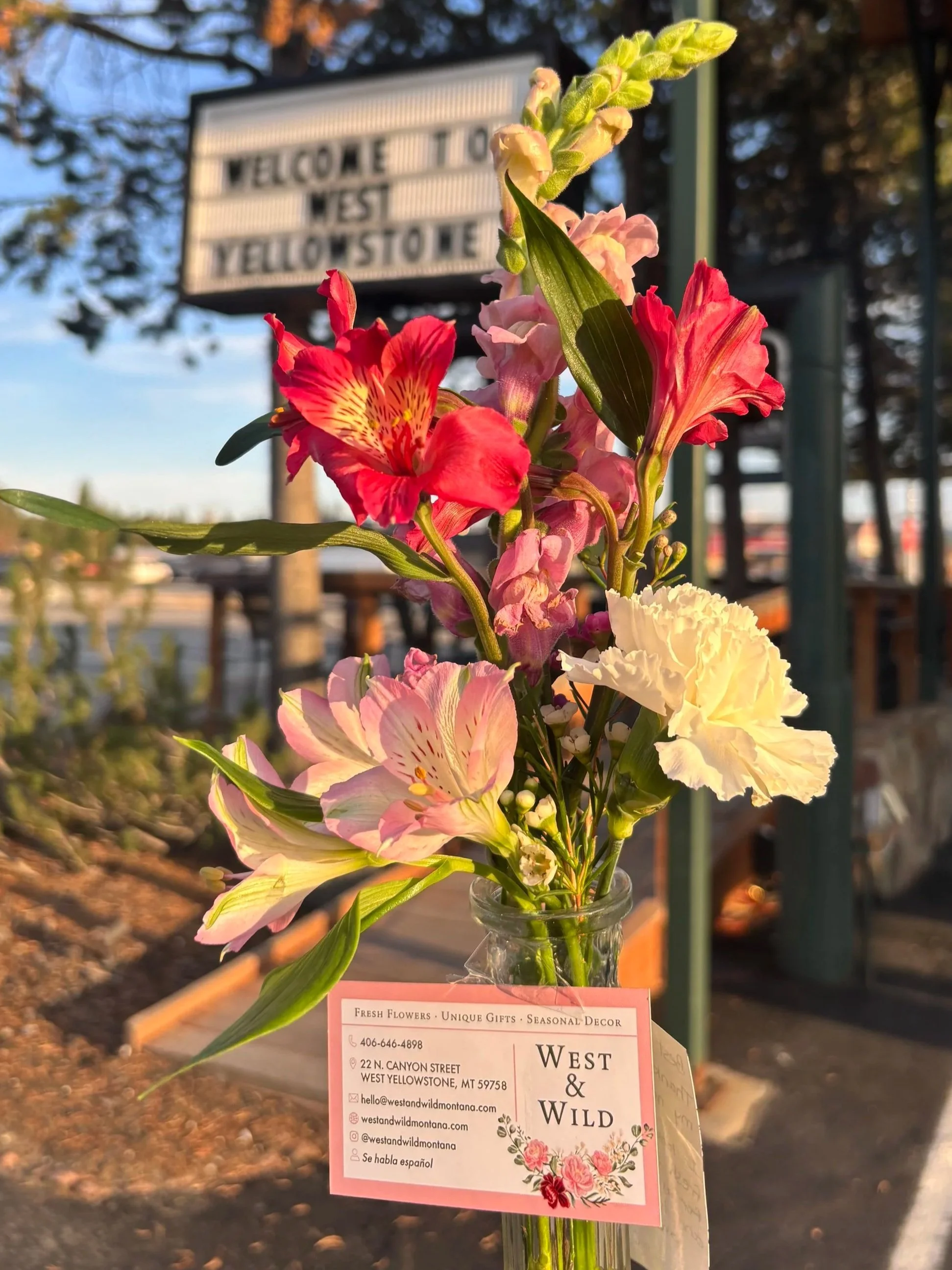 A bouquet of pink, red, and white flowers in a glass vase held in front of a sign that reads 'Welcome to West Yellowstone.'