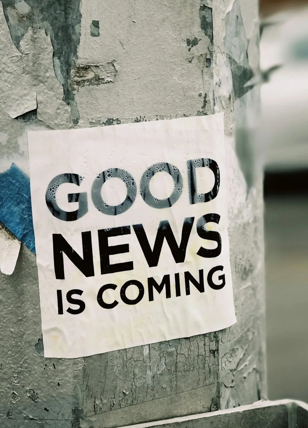 A torn paper poster on a weathered wall with the text 'Good News is Coming' with bubbles in the letters.