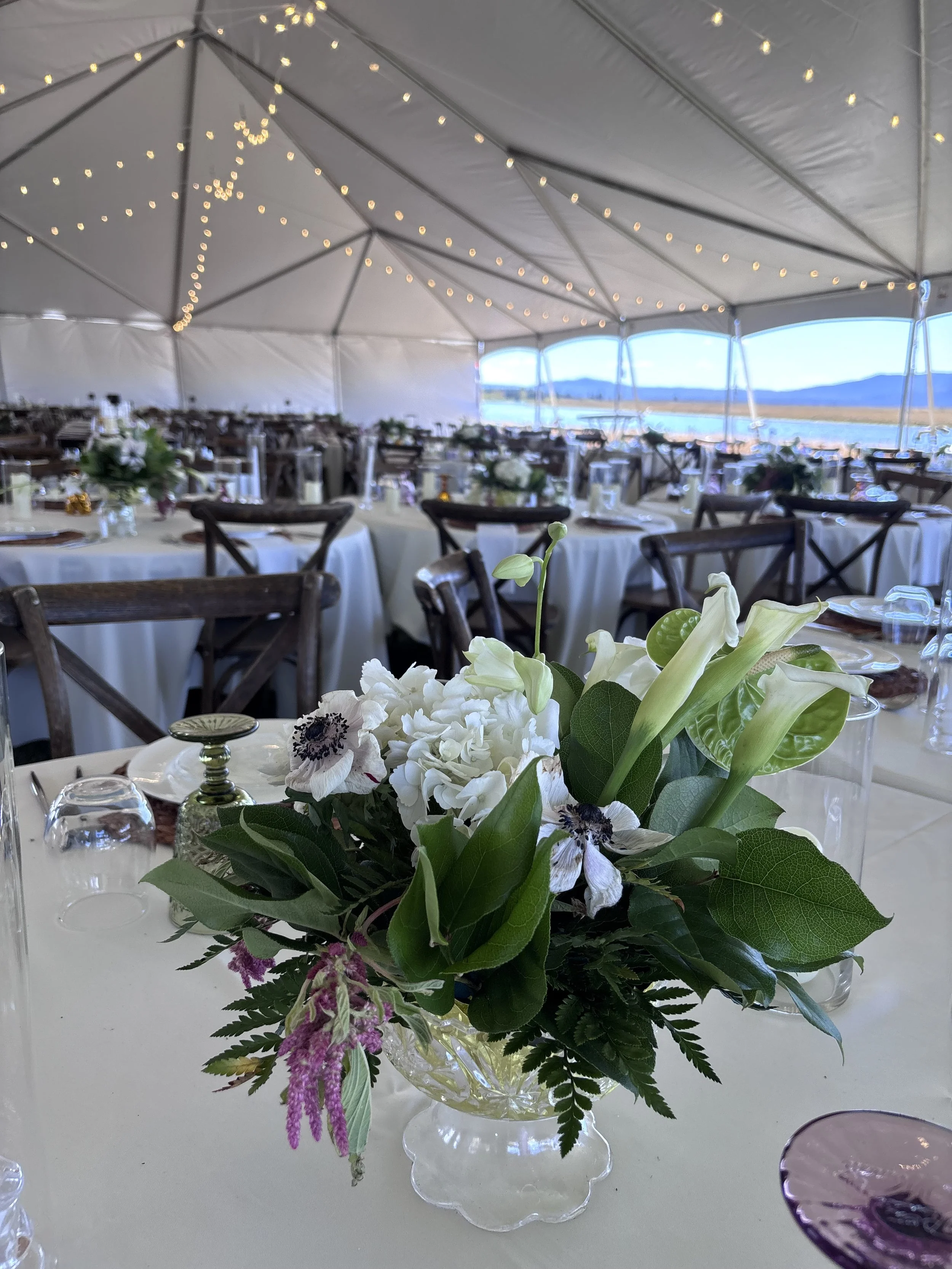 Flower arrangement with white flowers, green leaves, purple flowers, in a glass vase on a white table, at an outdoor event under a large tent with string lights, overlooking a body of water and mountains in the background.