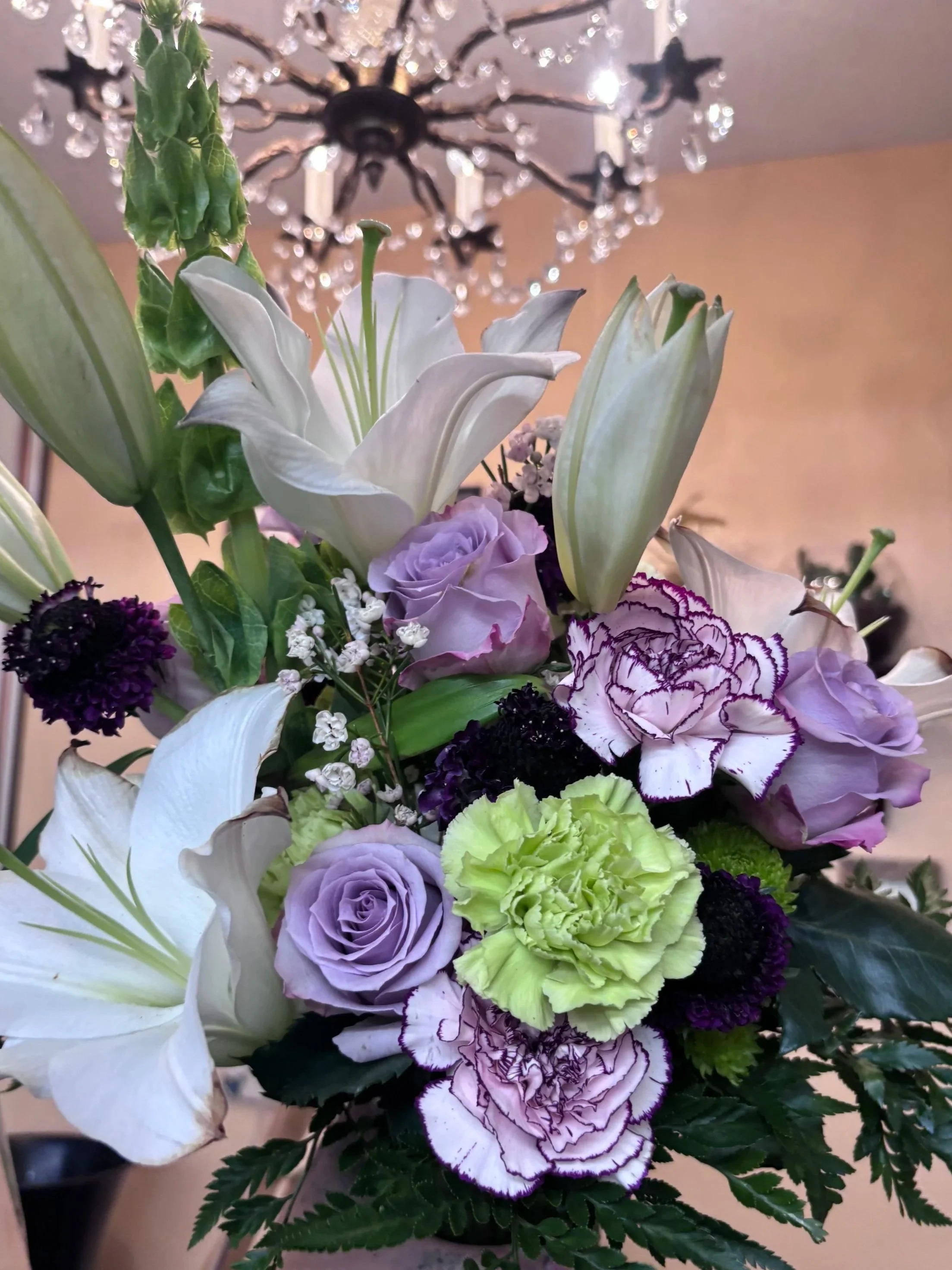 A close-up of a floral arrangement featuring white lilies, purple roses, purple and white striped carnations, light green carnations, and other small flowers, with a chandelier in the background.
