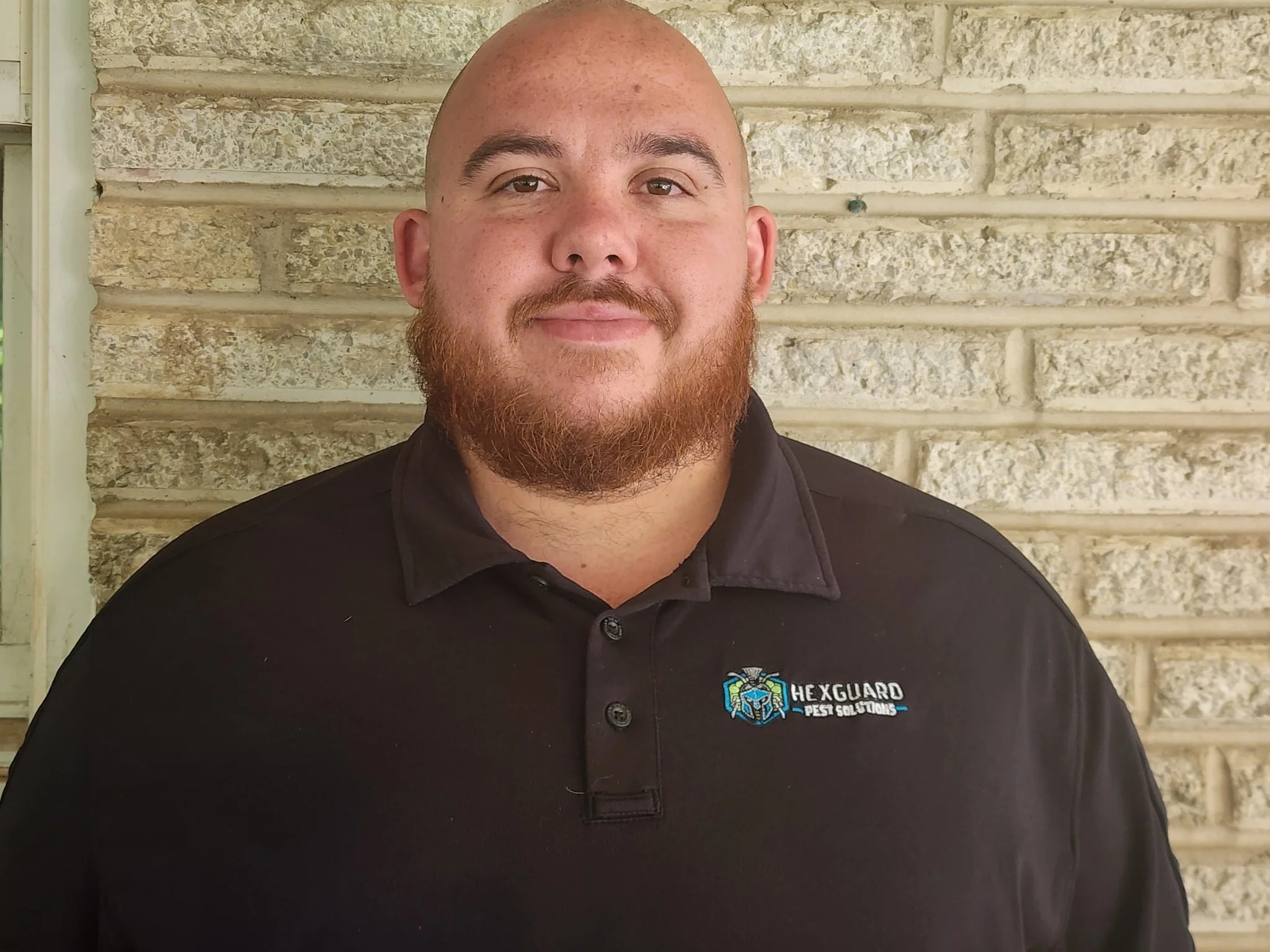 A man with a shaved head and a beard wearing a black collared shirt with an embroidered patch that reads "HEXGUARD PEST SOLUTIONS". He is standing in front of a brick wall.