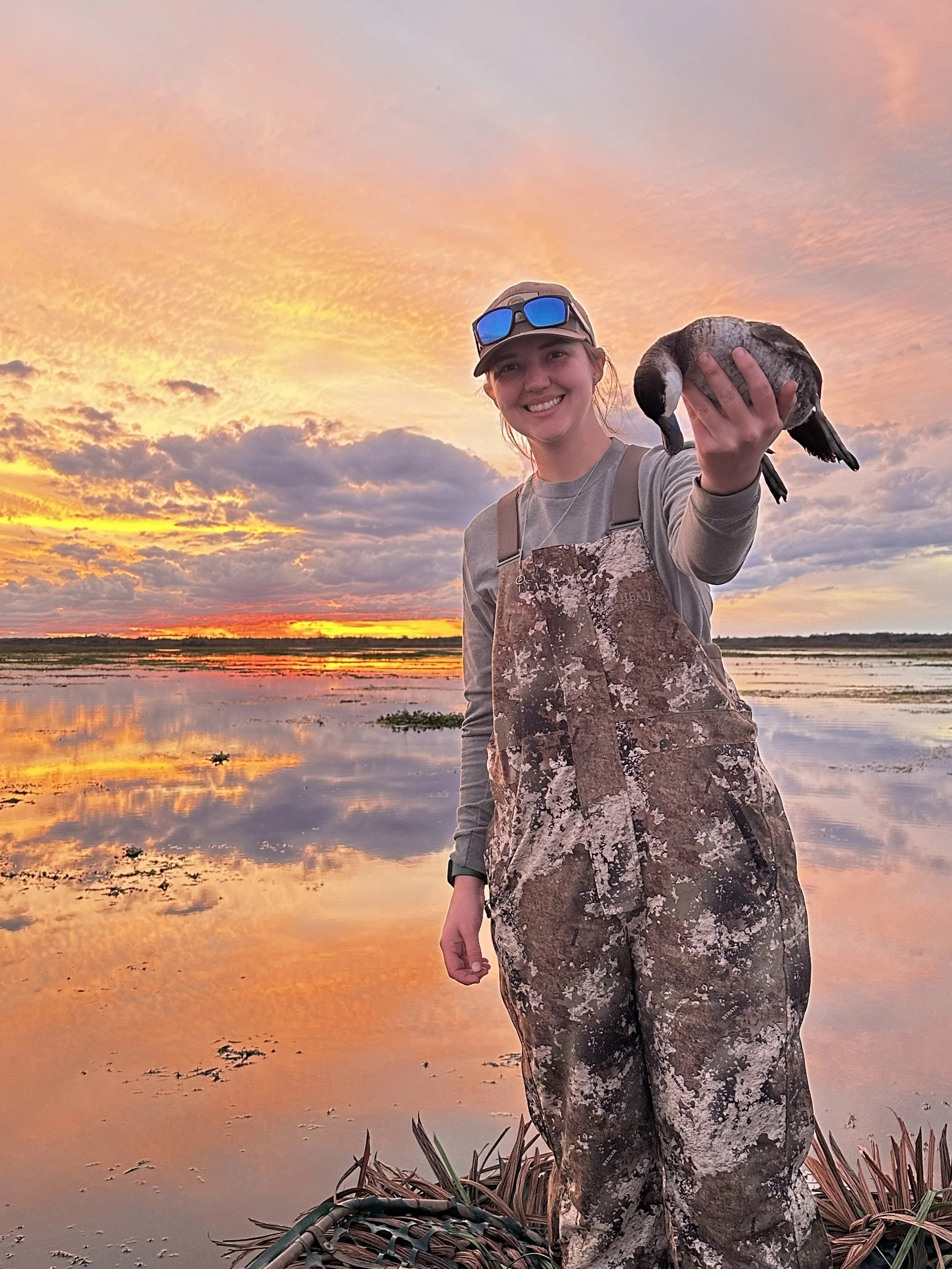 A woman dressed in camouflage clothing smiling and holding a duck near a body of water at sunset, with a vibrant orange and pink sky and clouds.
