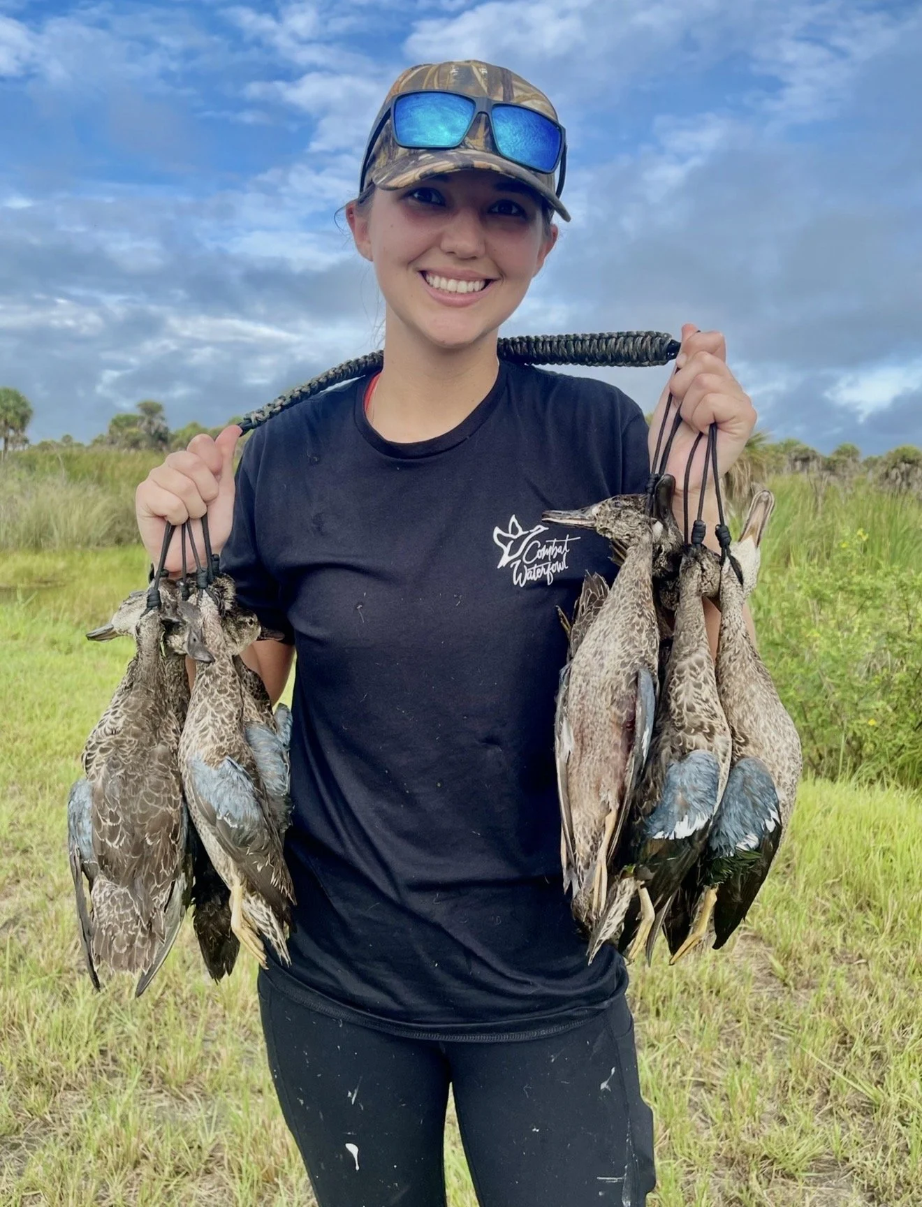 A young woman smiling outdoors in a grassy field under a cloudy sky, holding a stick with multiple ducks hanging from it.
