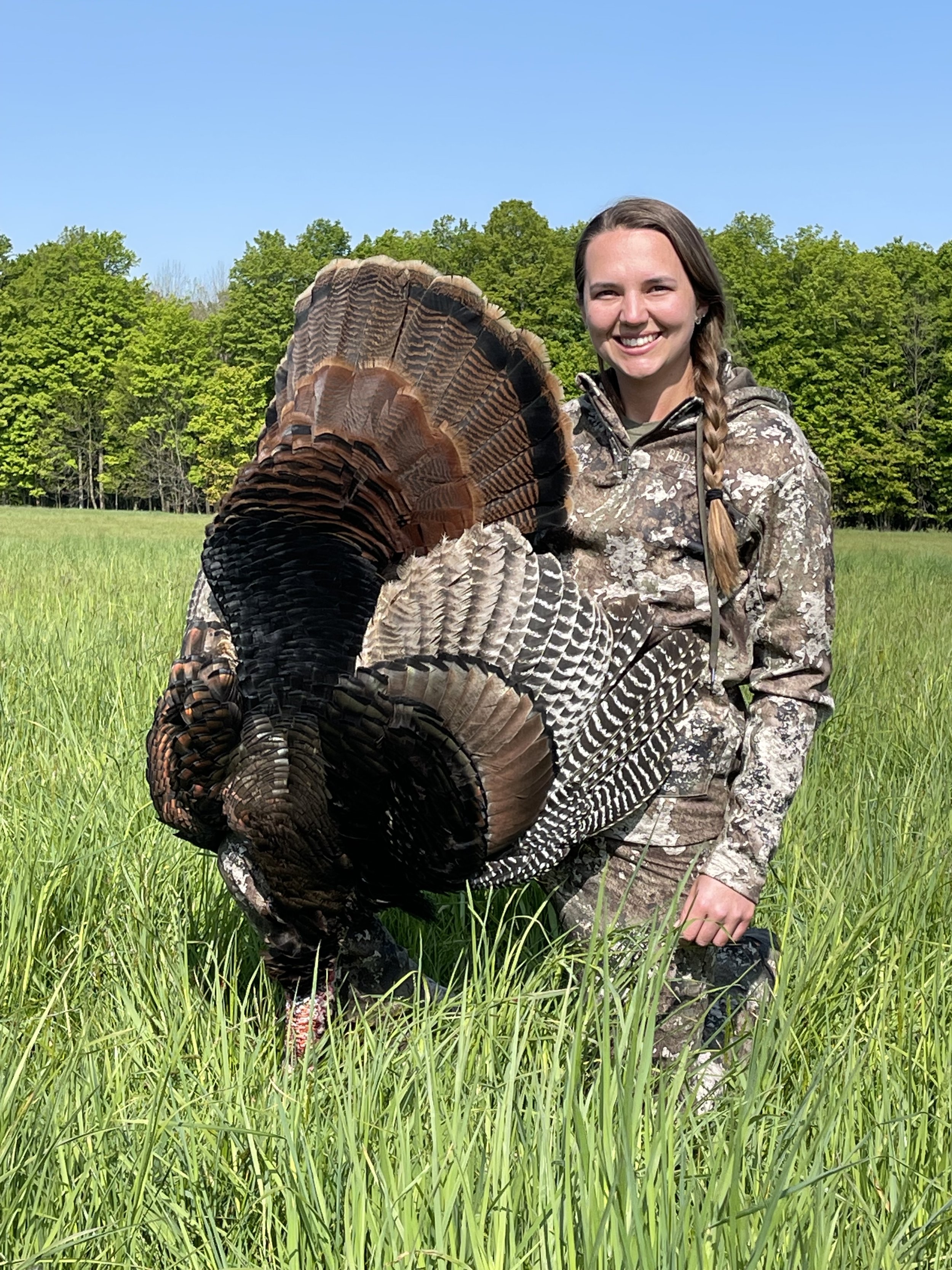 A woman in camouflage clothing smiling while kneeling in a grassy field, holding a large wild turkey with spread tail feathers.
