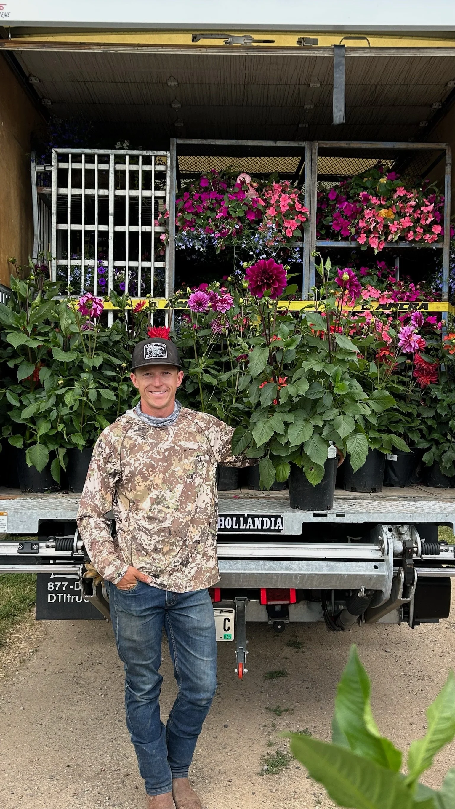 A man in camouflage clothing and a baseball cap stands in front of a truck loaded with pink flowering plants, with a backdrop of greenery.