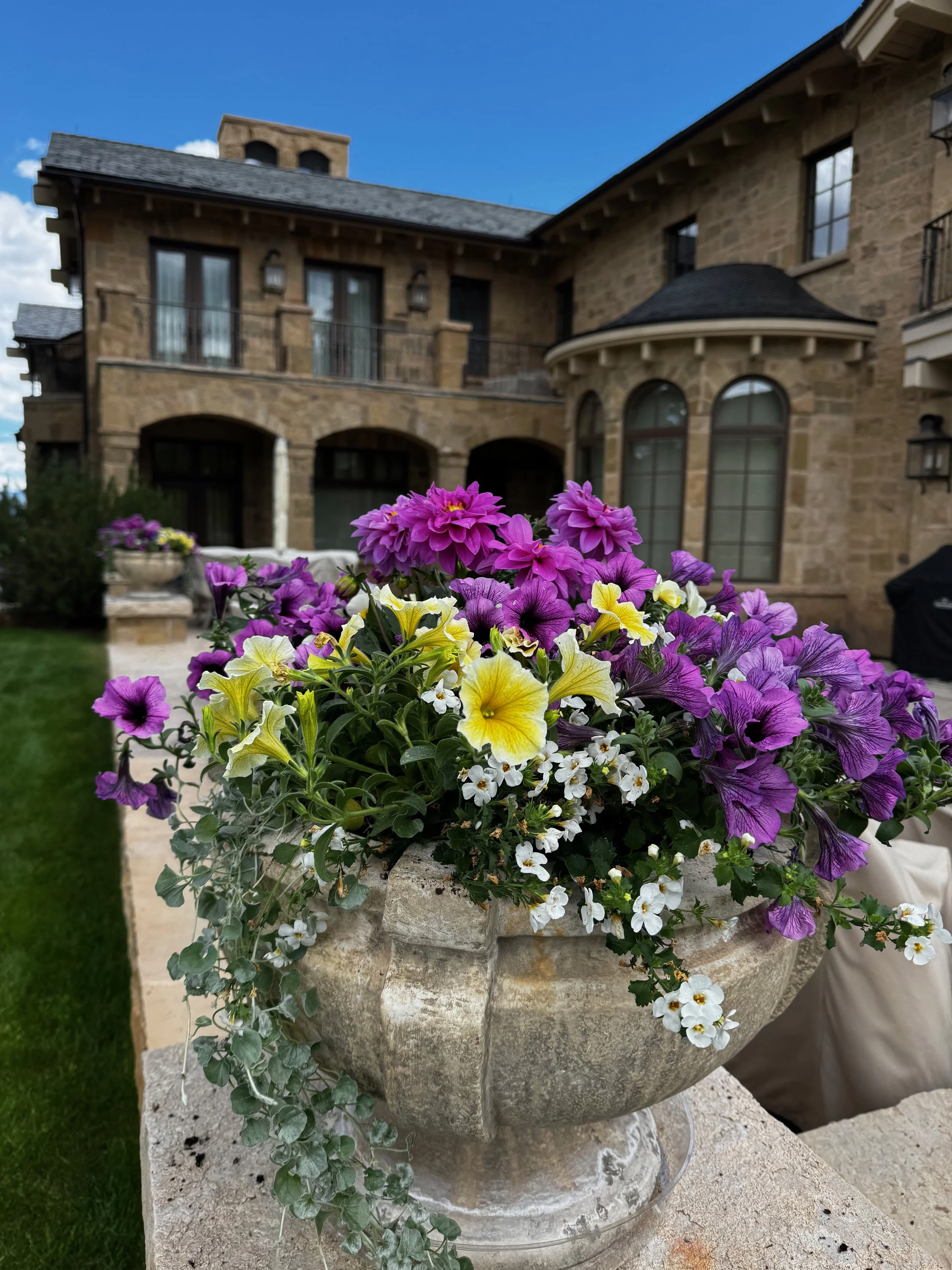 A large stone planter filled with purple, yellow, white, and violet petunias and other flowers, placed on a stone surface outside a stone house with multiple windows and a balcony under a blue sky.