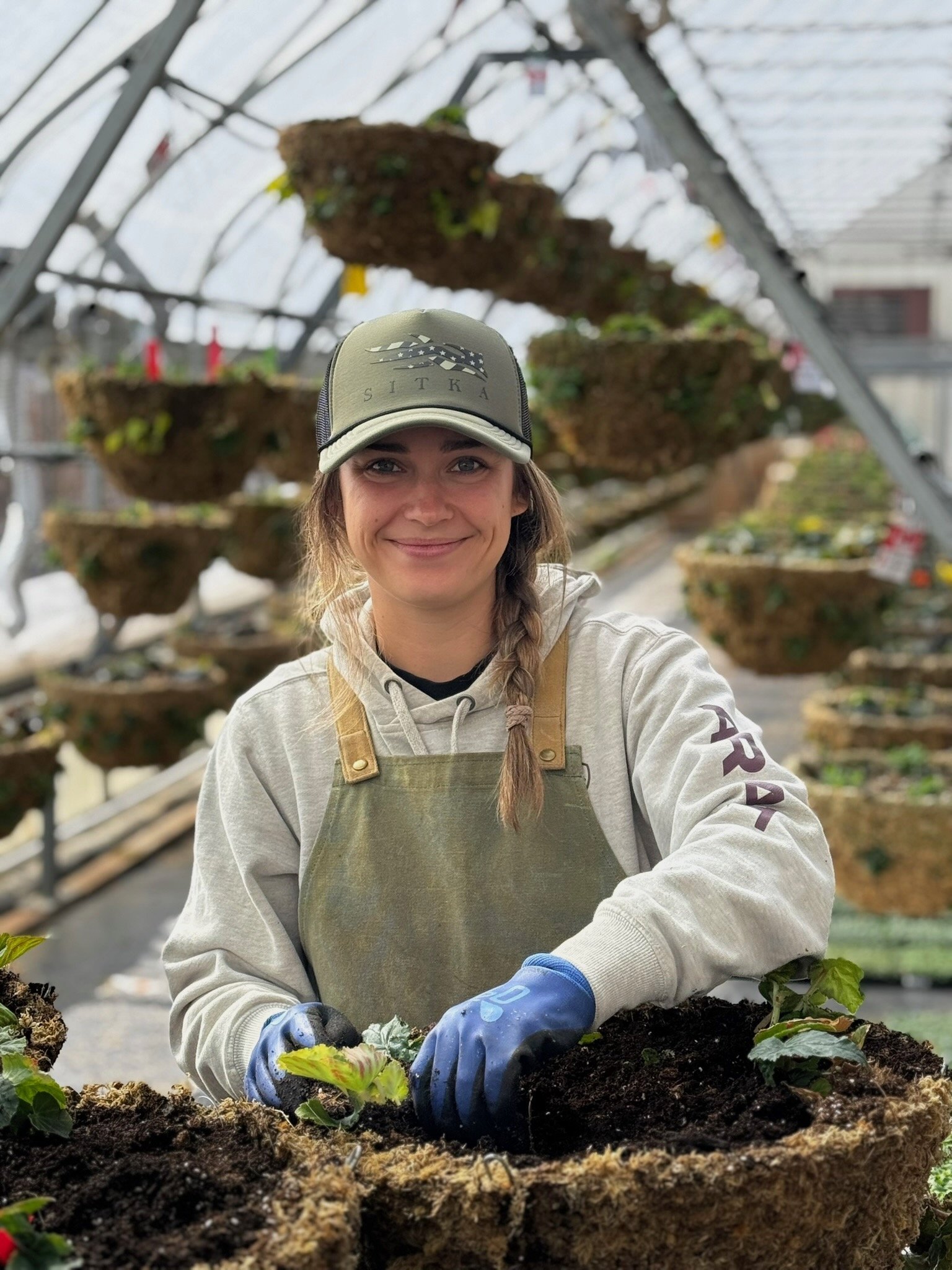 A woman wearing a gray hoodie, green apron, and blue gloves is gardening inside a greenhouse. She is planting or tending to young plants, with a metal structure and hanging planting beds filled with soil and plants visible in the background.