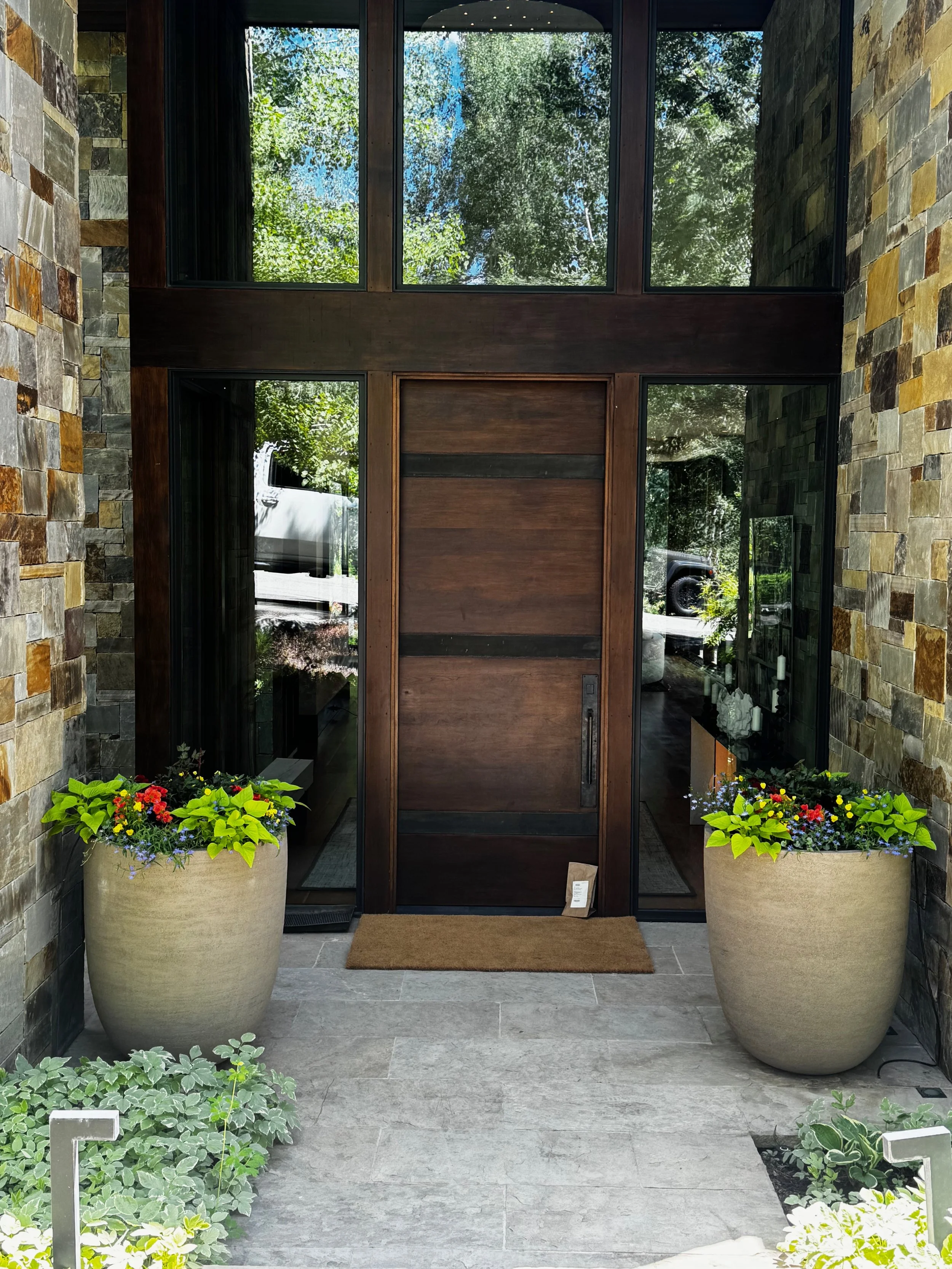 A modern entryway with a wooden door and large glass side panels, flanked by two large beige planters with colorful flowers, with a stone wall on both sides and green trees visible through the glass.