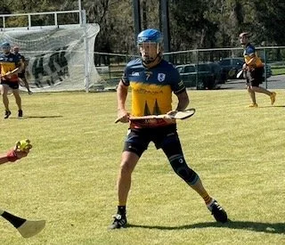 Lacrosse player in a blue and yellow uniform holding a lacrosse stick on a grass field with other players in the background.