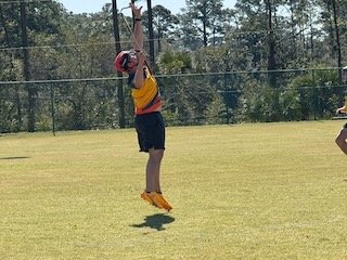 Child wearing a helmet and sports uniform hanging from a horizontal bar on a grassy field.
