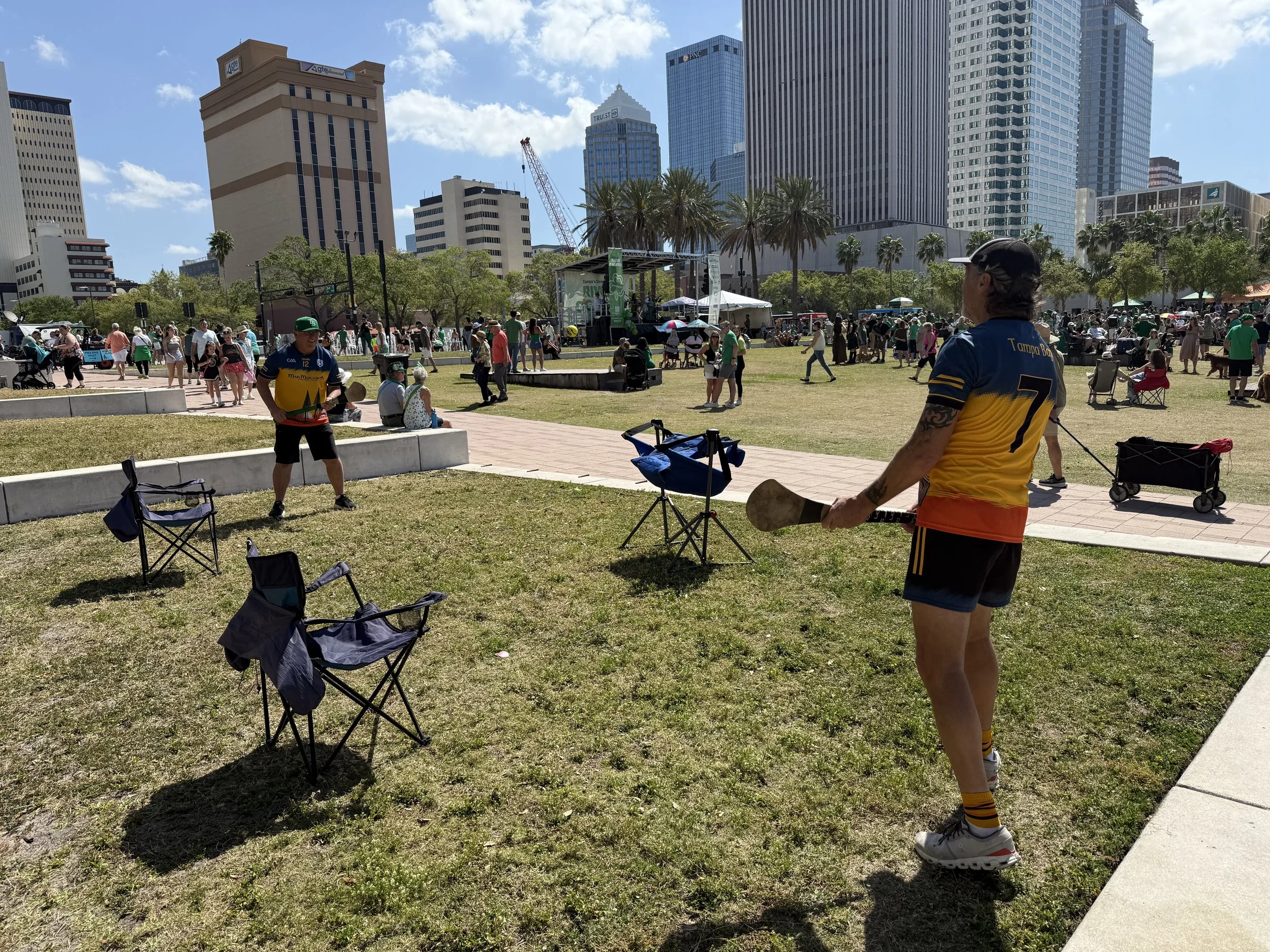 People gather in an outdoor park area in a city with tall skyscrapers. Some people are sitting, others are walking or standing. There are tents, umbrellas, and a stage set up, indicating a festival or outdoor event. The weather is sunny with a blue s