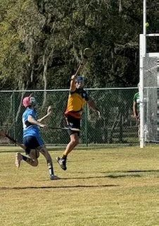 Two children playing football on a grassy field with trees and a chain-link fence in the background.