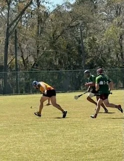 Lacrosse players on a grassy field during a game, with trees and a fence in the background.