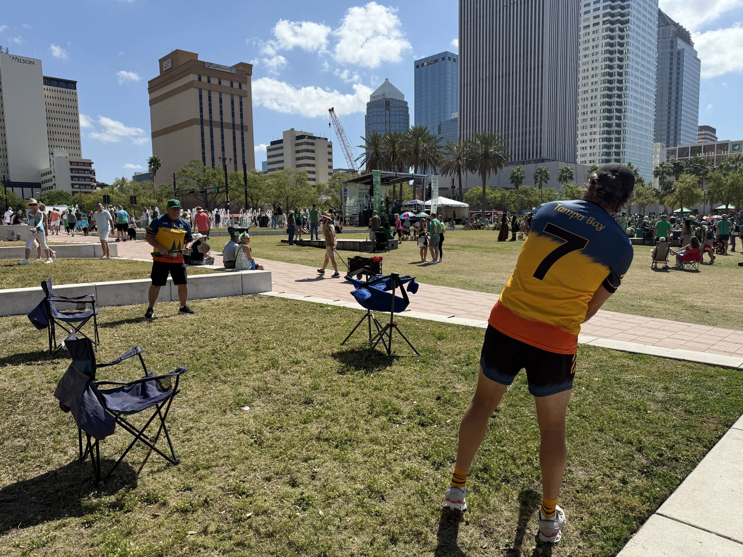A scene in an urban park during a sunny day with many people, including a man in a Tampa Bay Buccaneers jersey, nearby chairs, and a city skyline with tall buildings in the background.