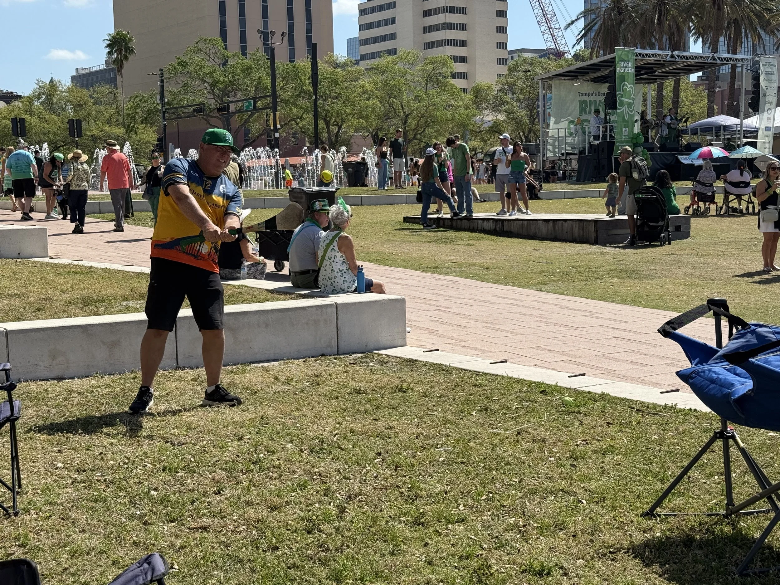 Outdoor concert event in an urban park with a stage, musicians, and audience, surrounded by trees and tall buildings in the background.
