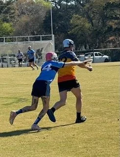 Two young players in a football game, one in a blue jersey and the other in a multicolored jersey, with the player in multicolored jersey holding the football and running while the player in blue tries to tackle.