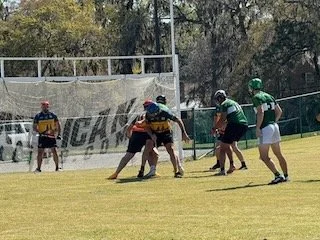 Two individuals in a game of lacrosse, with one player in black and yellow and the other in green and white, on a grassy field in an outdoor setting, with other players and a coach nearby.