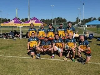 Group of nine men in yellow and black sports uniforms posing on a grassy field with tents and trees in the background.