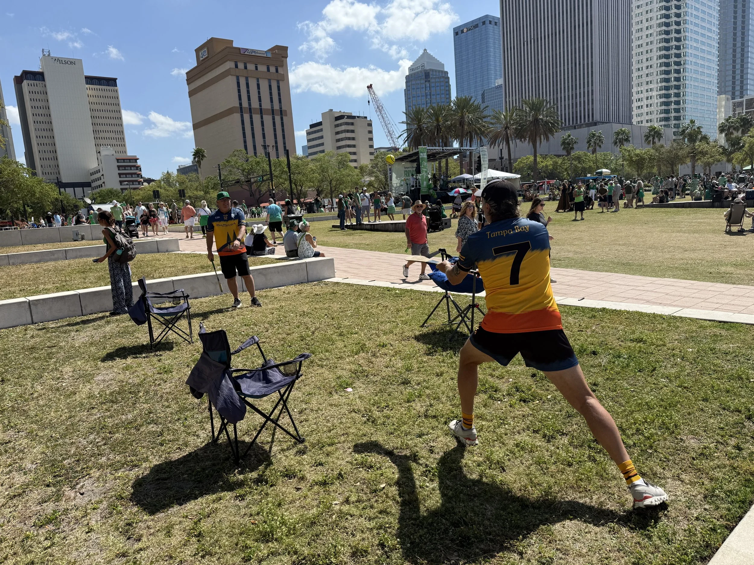 People gathering in a park with high-rise buildings in the background. Some are sitting, walking, and participating in an event near a stage, with palm trees lining the area on a sunny day.