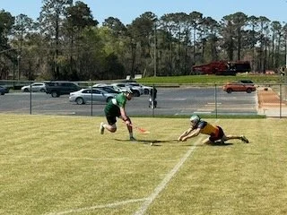 Two young men playing rugby on a grass field, one reaching for the ball while the other is on the ground. A parking lot with cars and trees in the background.