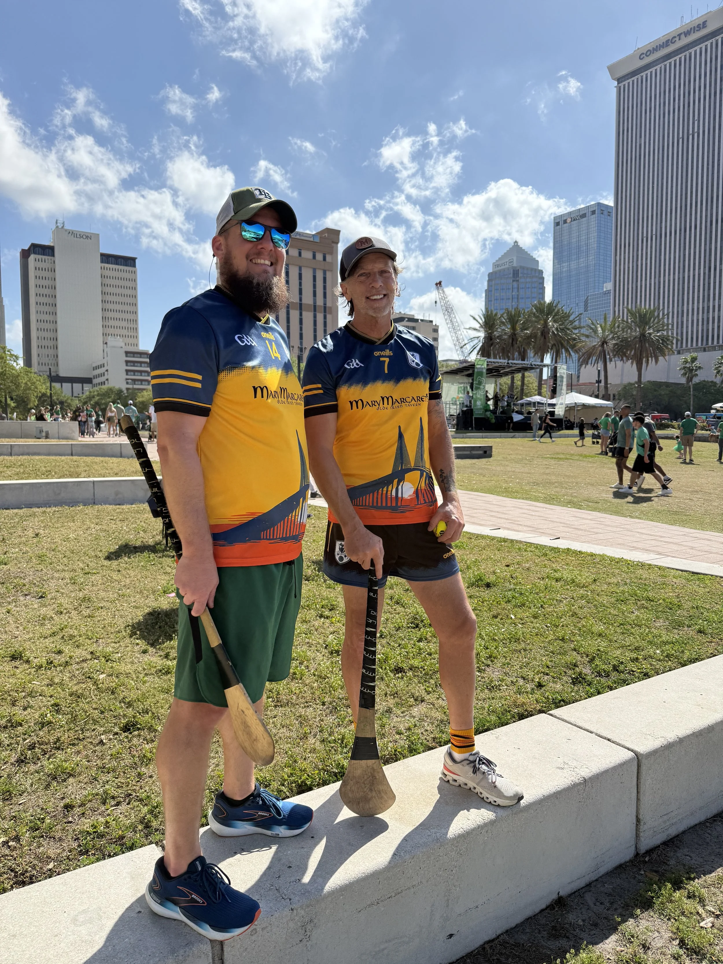 Two men smiling and holding wooden paddles in an urban park with tall buildings, palm trees, and a stage with tents in the background on a sunny day.