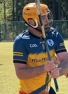 Person wearing a yellow helmet, blue and yellow sports jersey, holding a hurling stick on a field.